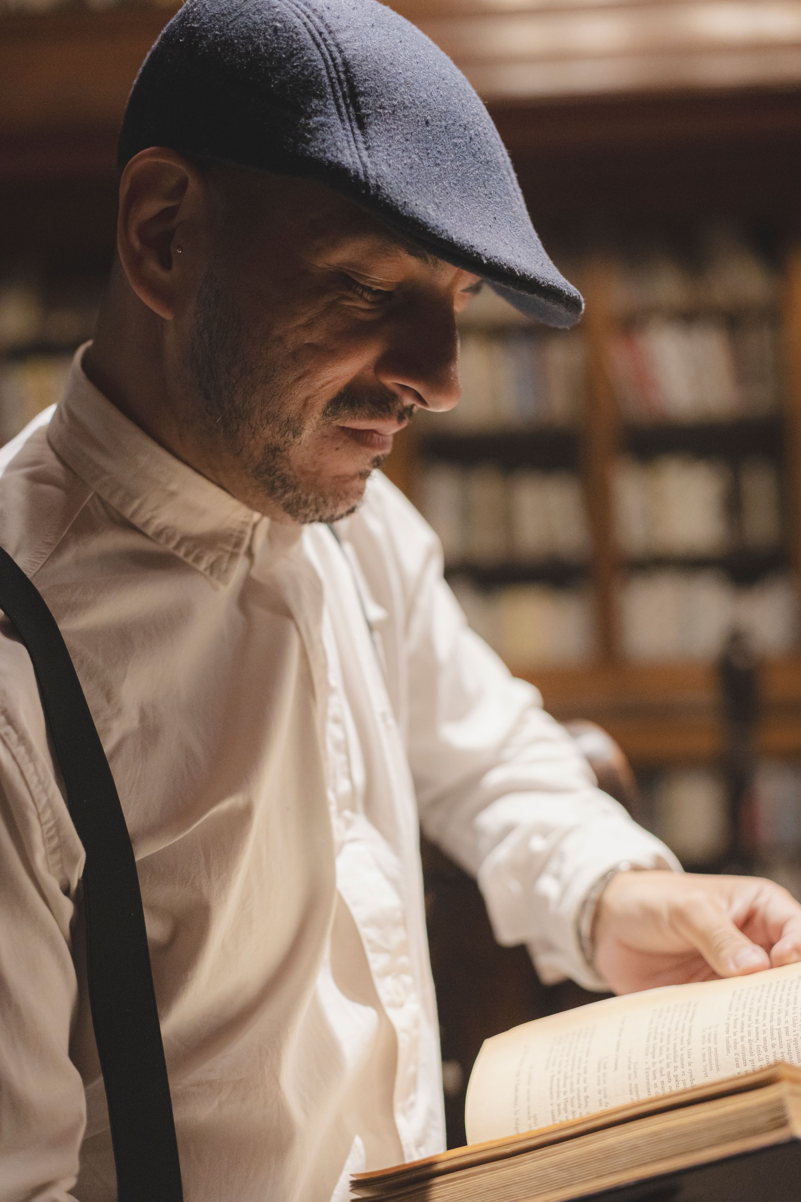A man in a stylish hat reading a book in a warm, inviting library.