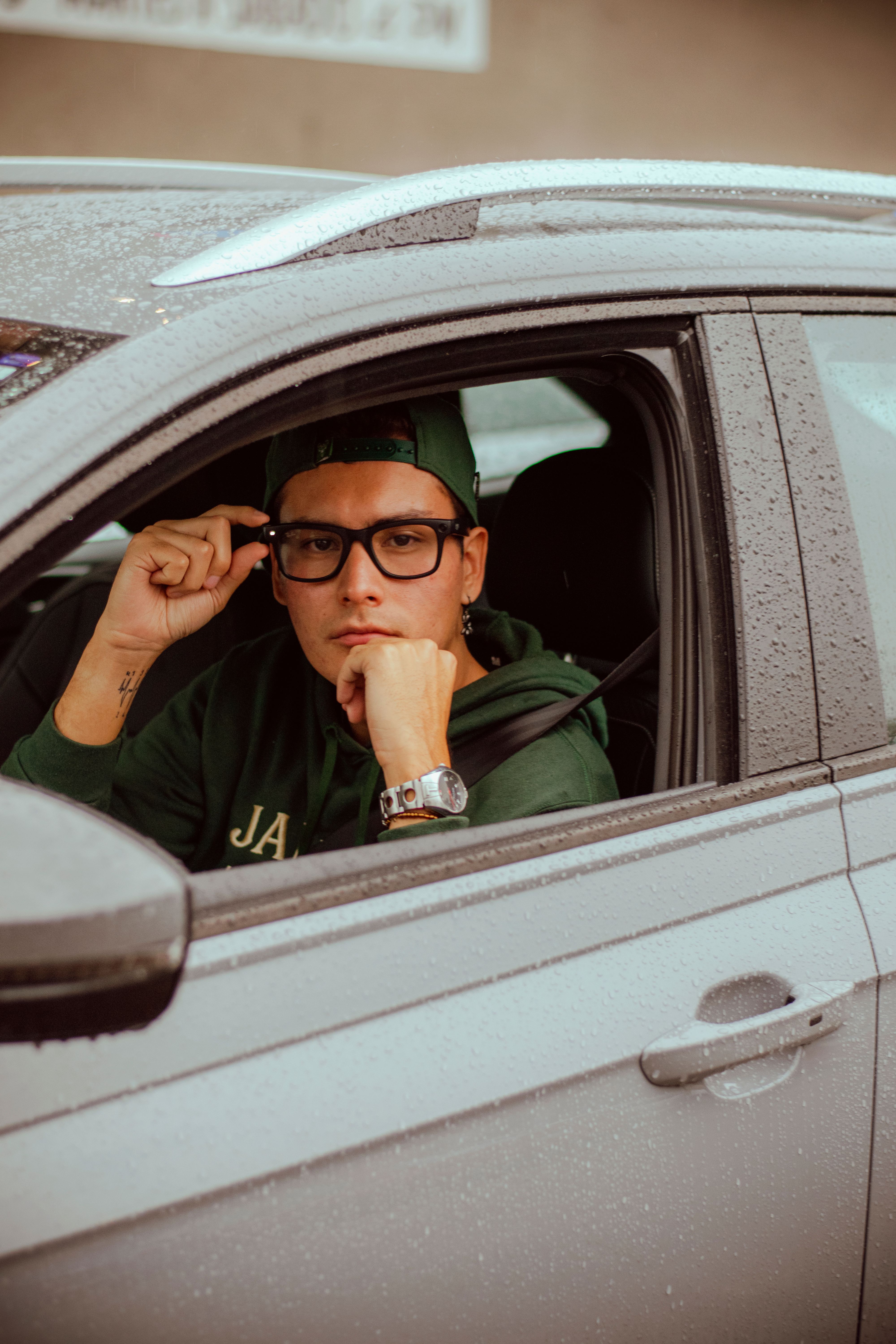 Young man in glasses sitting in car on rainy day, looking outside the window.