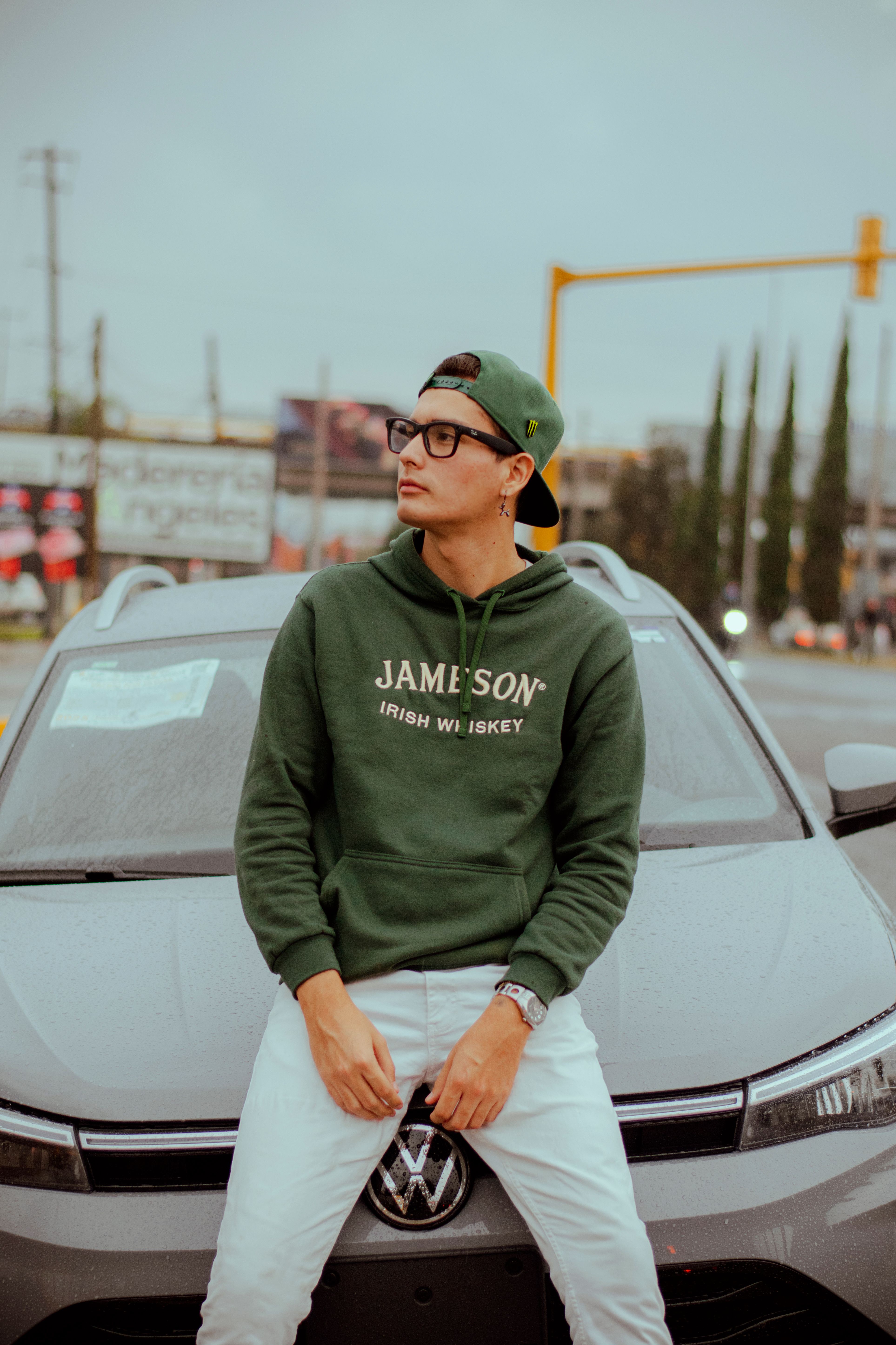 Young man sitting on car hood, wearing green hoodie and cap in urban area.