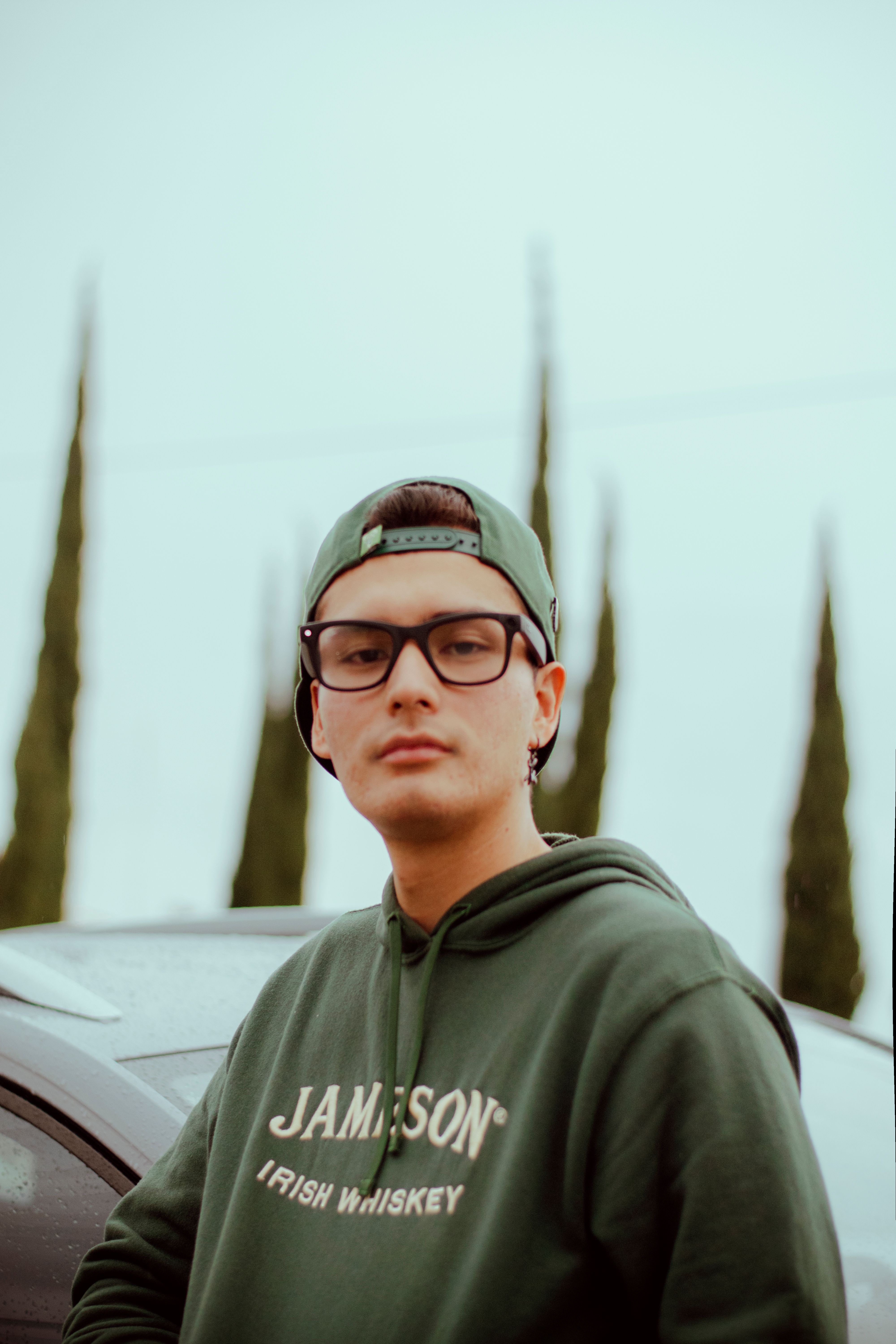 A young man wearing glasses and a green hoodie stands confidently beside a car.