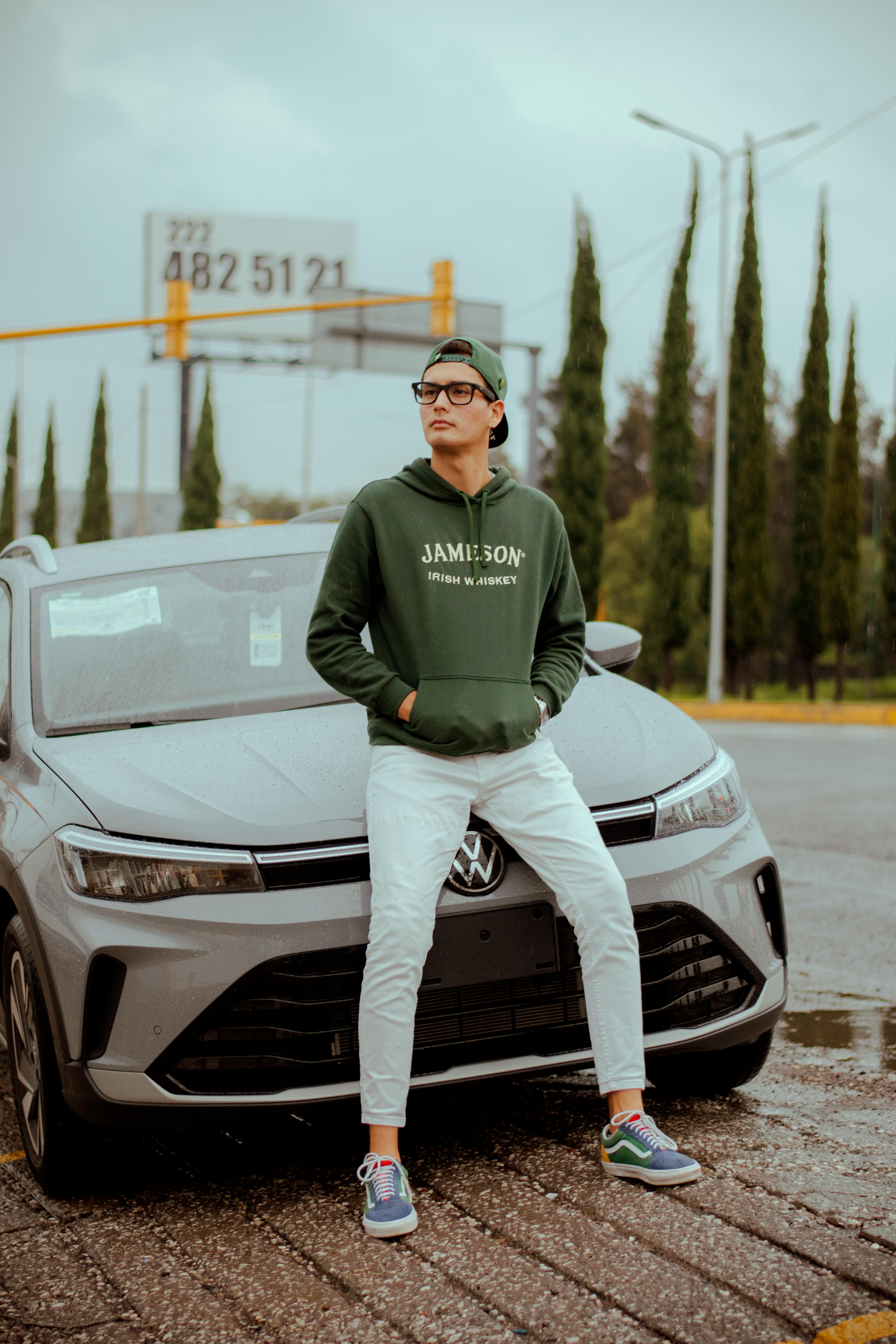 Young man in casual wear leaning on car outdoors, urban vibe.