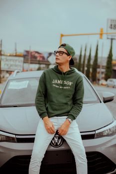 Stylish young man in green hoodie and glasses poses on a car hood outdoors.