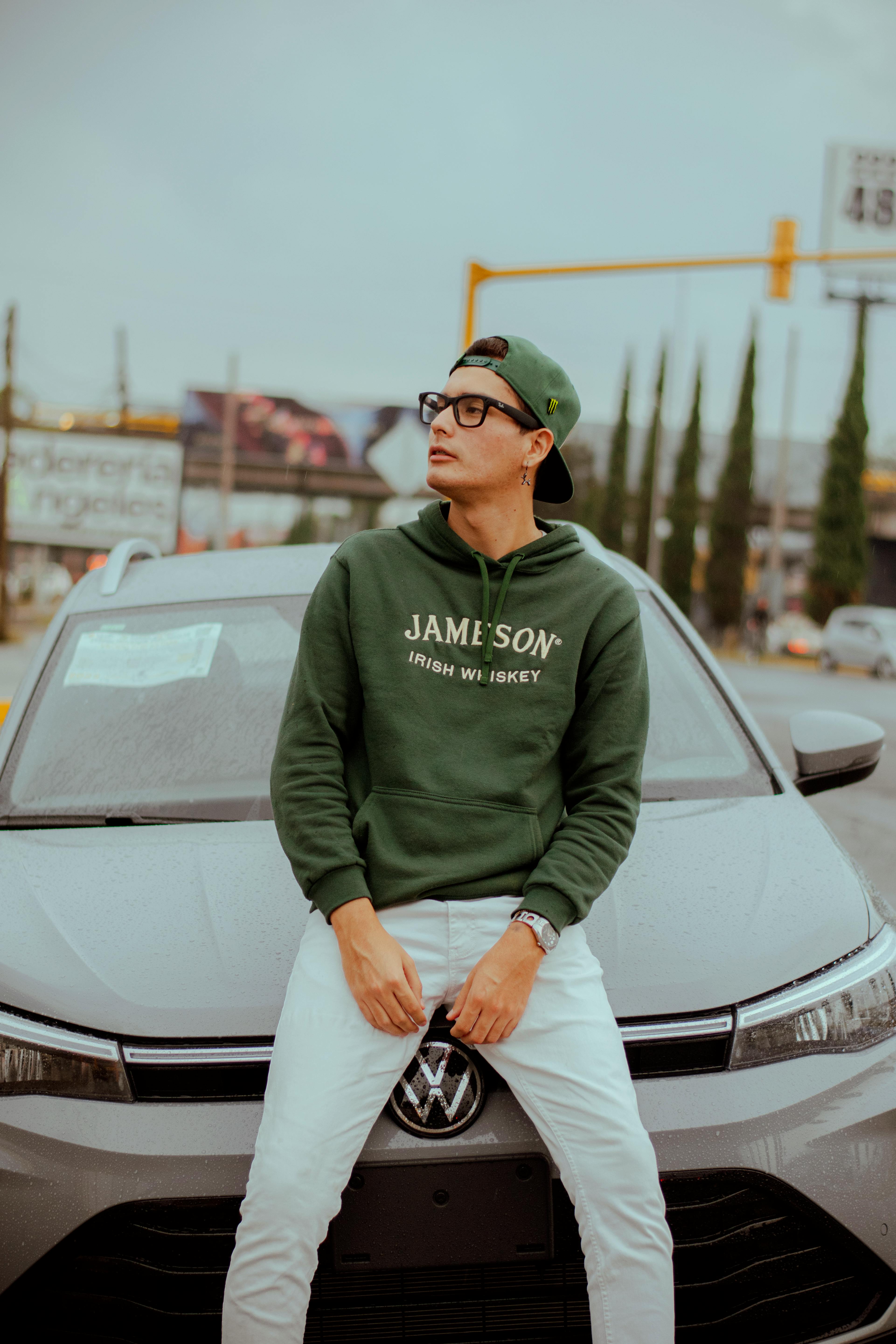 Free Stylish young man in green hoodie and glasses poses on a car hood outdoors. Stock Photo