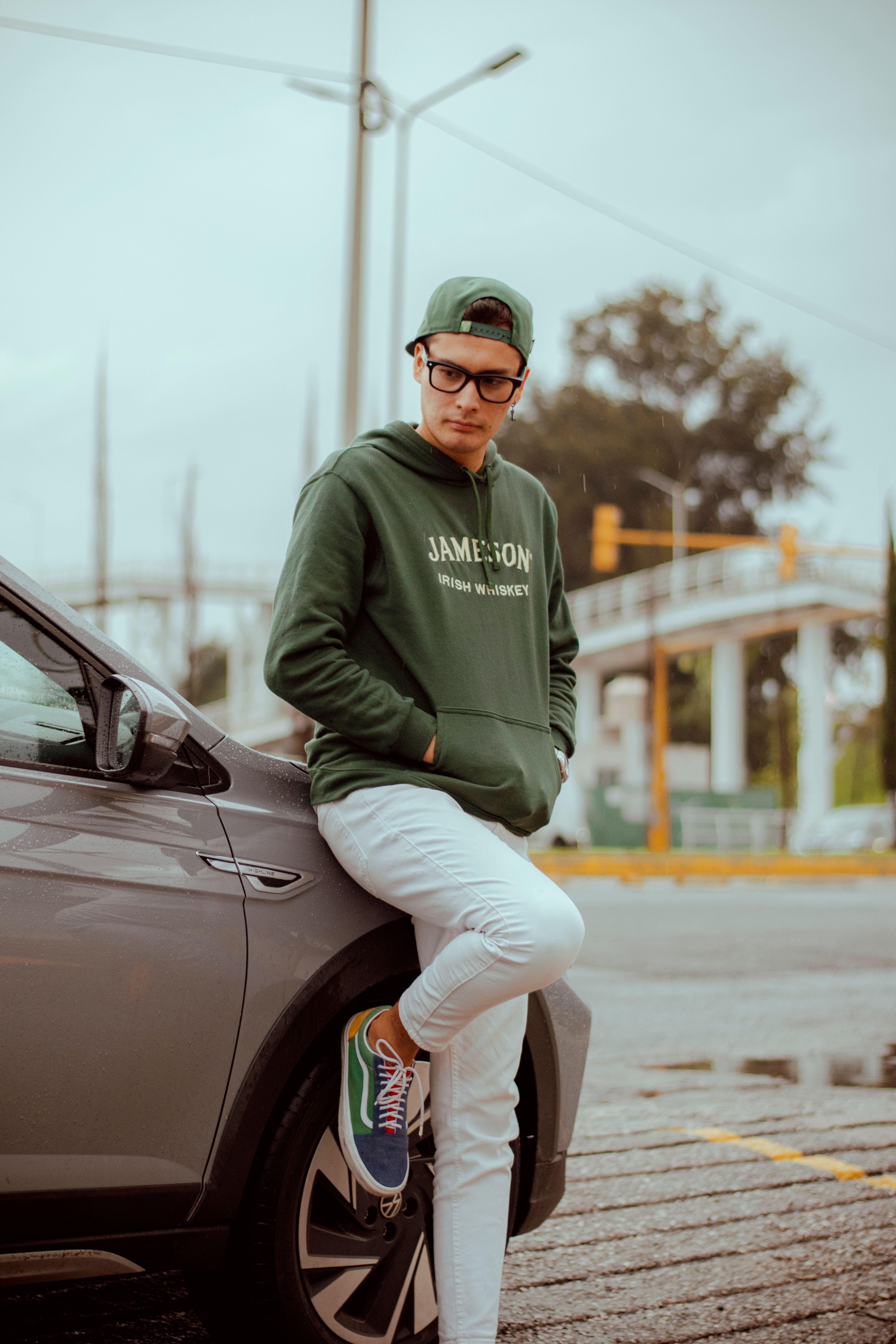 Casual young adult in green hoodie and cap leaning on a car, urban background.