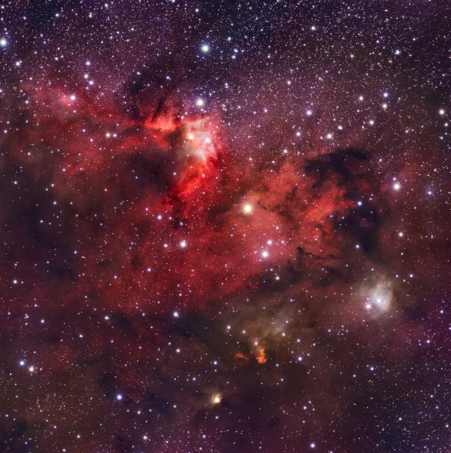 A red and orange emission nebula threaded with dark dust among dense stars