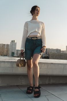 Fashionable woman in casual outfit posing on a rooftop during sunset in an urban setting.