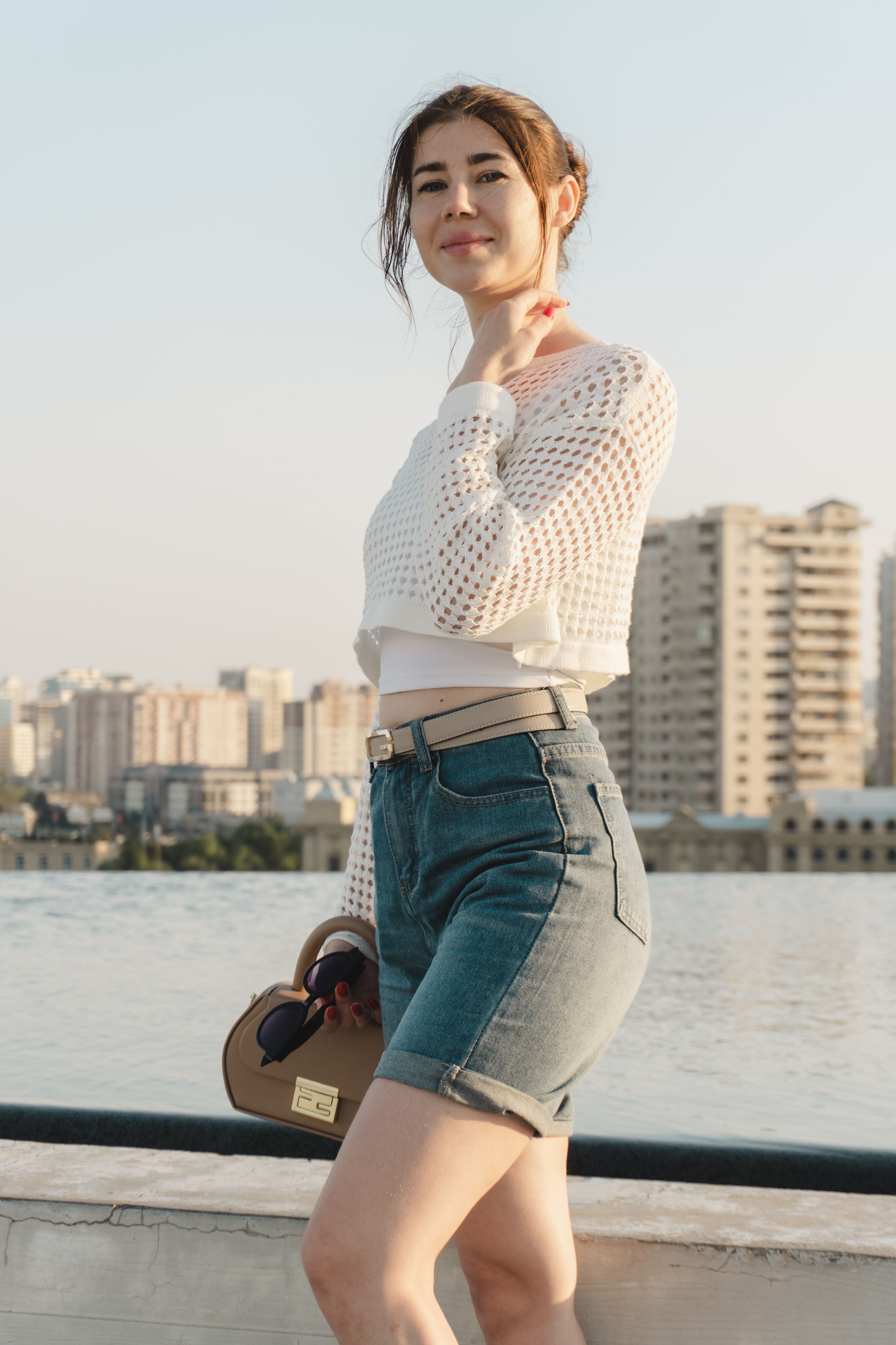 Uma mulher elegante em traje de verão posando perto da água com uma paisagem urbana ao fundo.