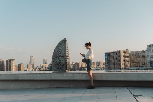 Young woman checking her phone on a rooftop with city skyline and modern architecture in the background.