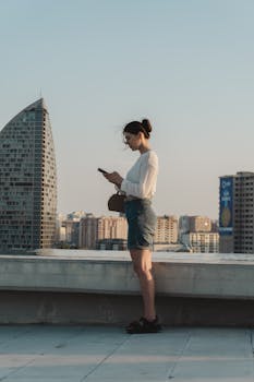 A woman stands on a rooftop texting with a city skyline in the background during the day.