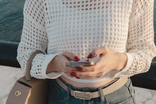 Free Close-up of a woman using a smartphone outdoors, wearing a white knitted top and jeans. Stock Photo
