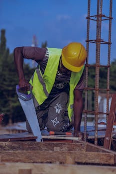 A construction worker in protective gear using a handsaw at a building site during the day.