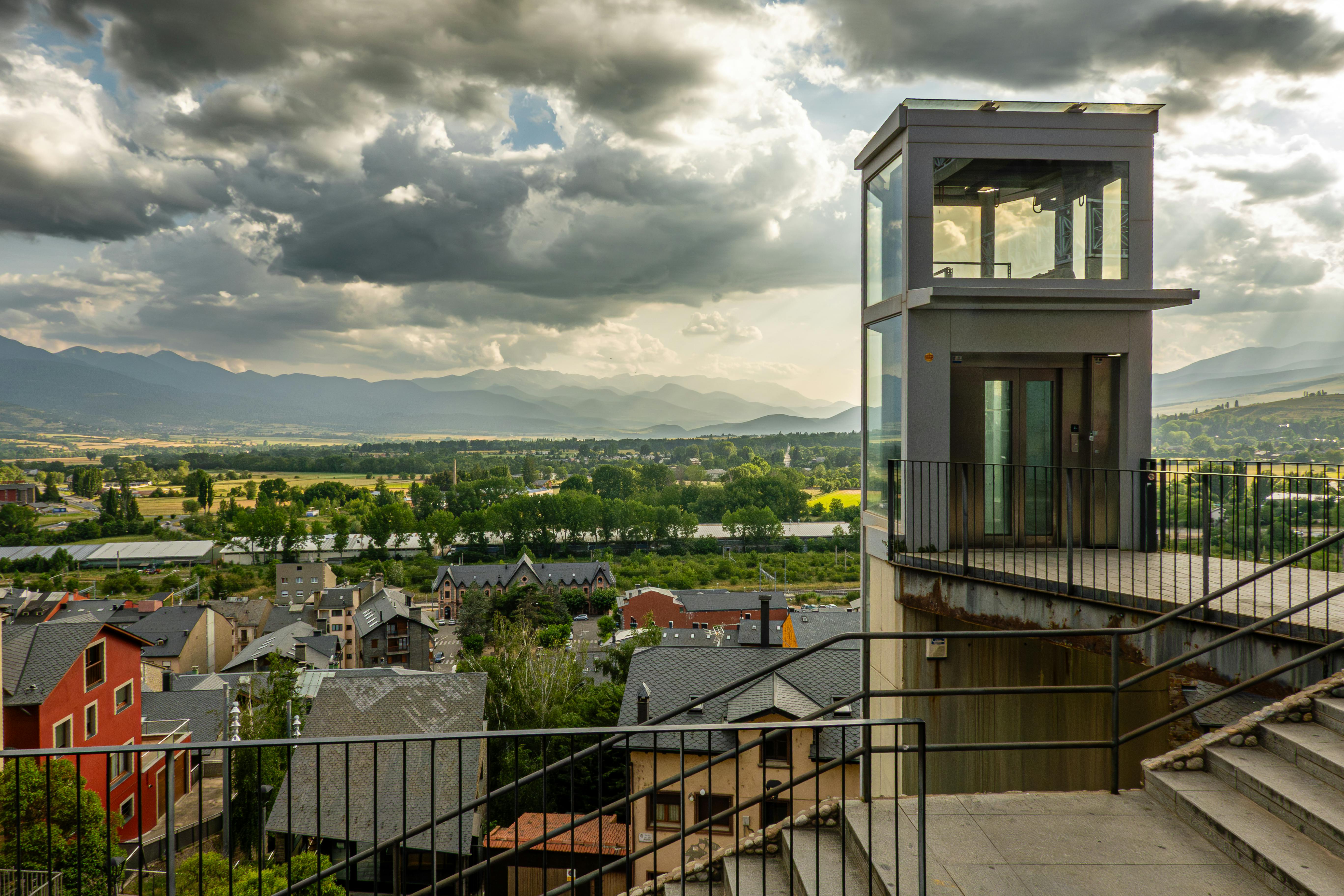 A modern elevator overlooks a picturesque town with mountains in the background under dramatic skies.