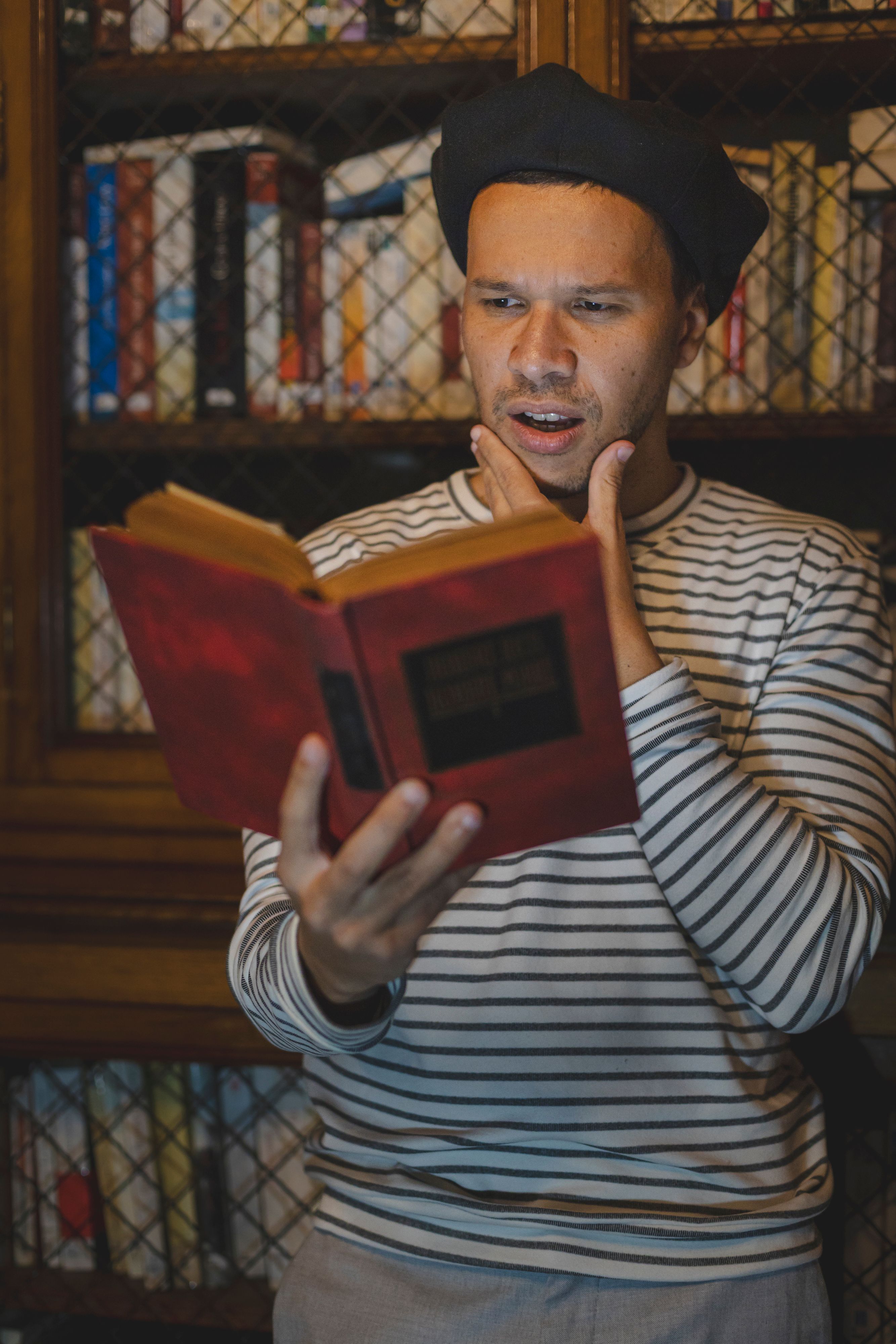 Man wearing a beret reading a book in a library with an expressive face.