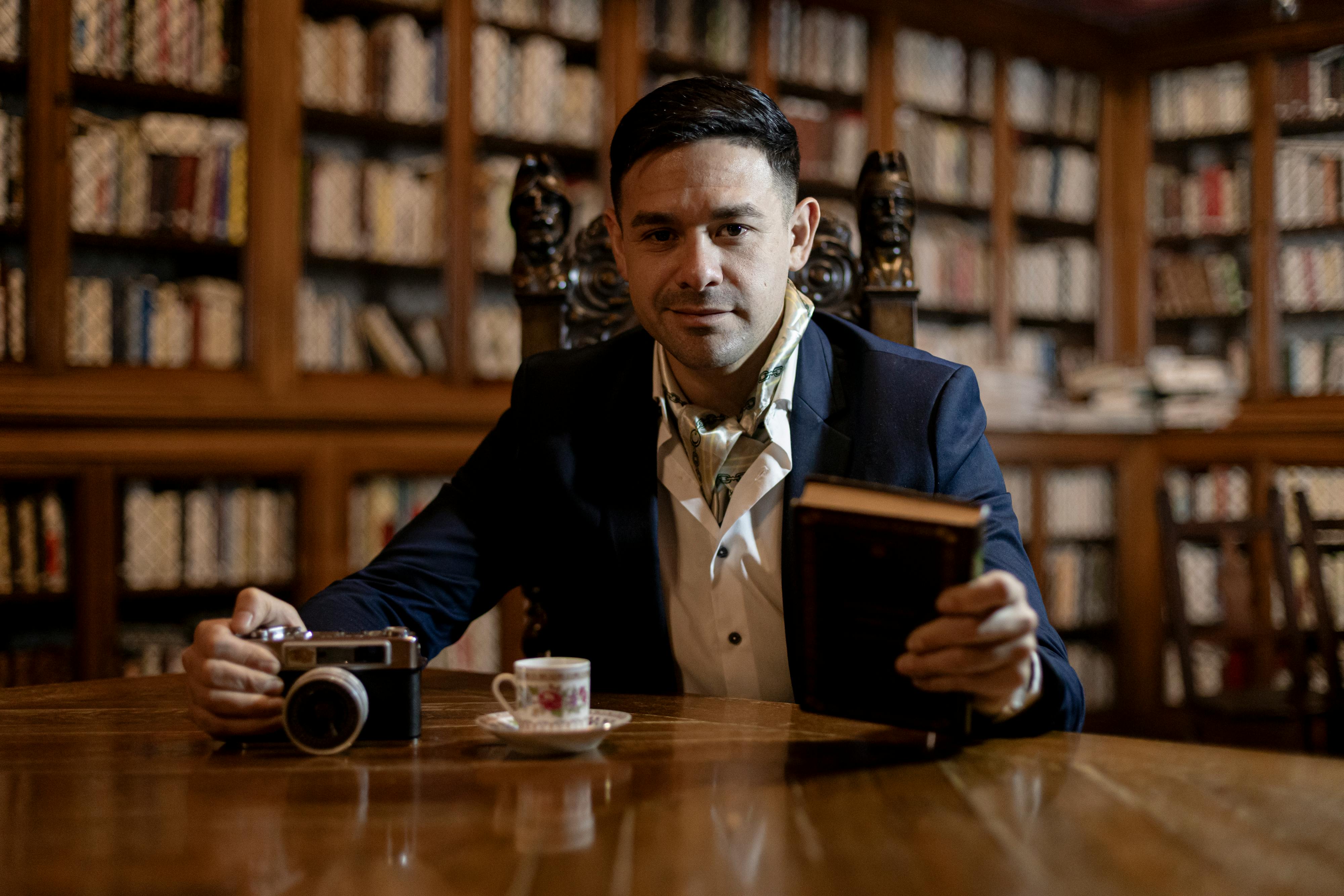 Man in a suit in a classic library holding a book and camera.