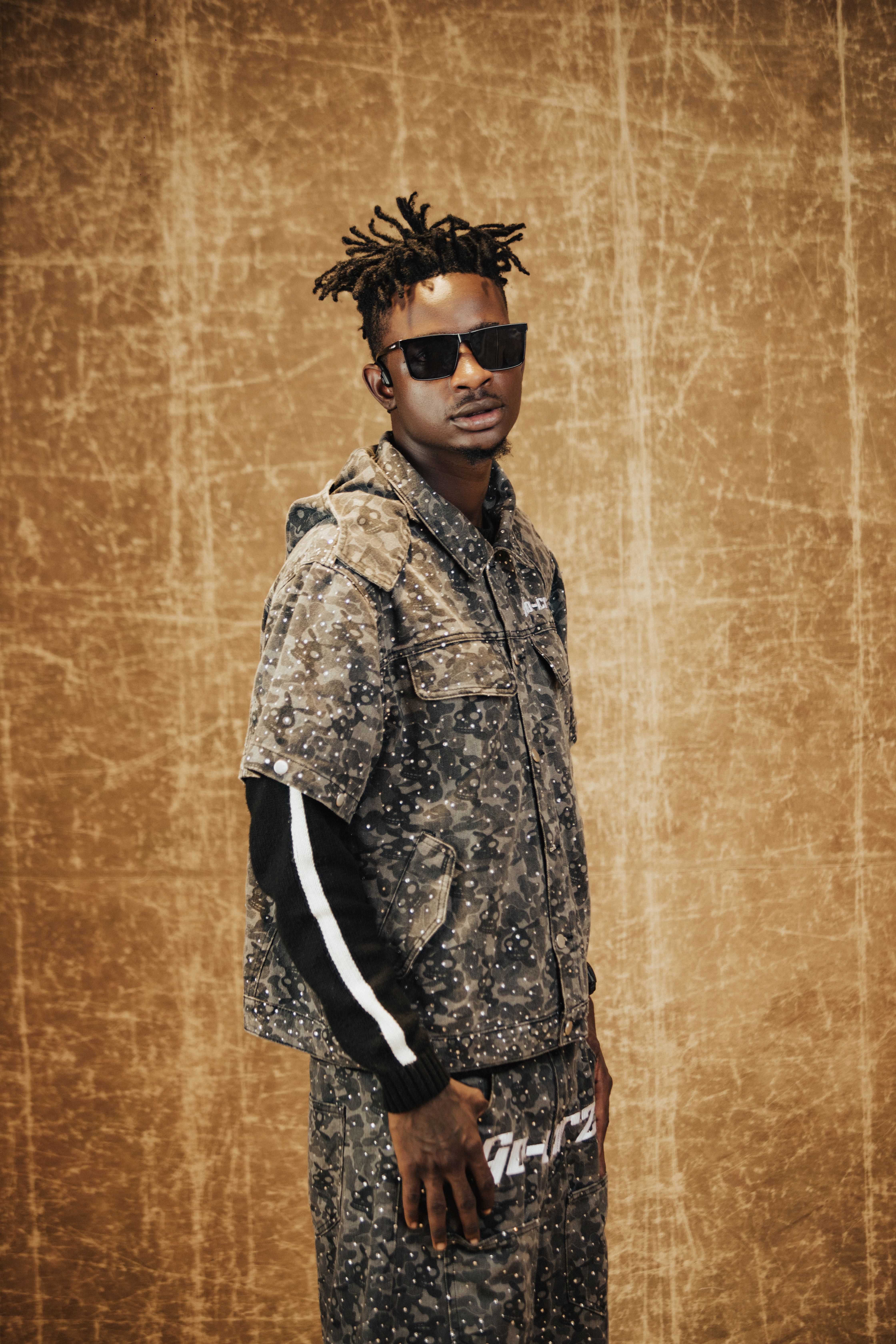 Portrait of a fashionable young man wearing a camouflage outfit and sunglasses against a textured background.