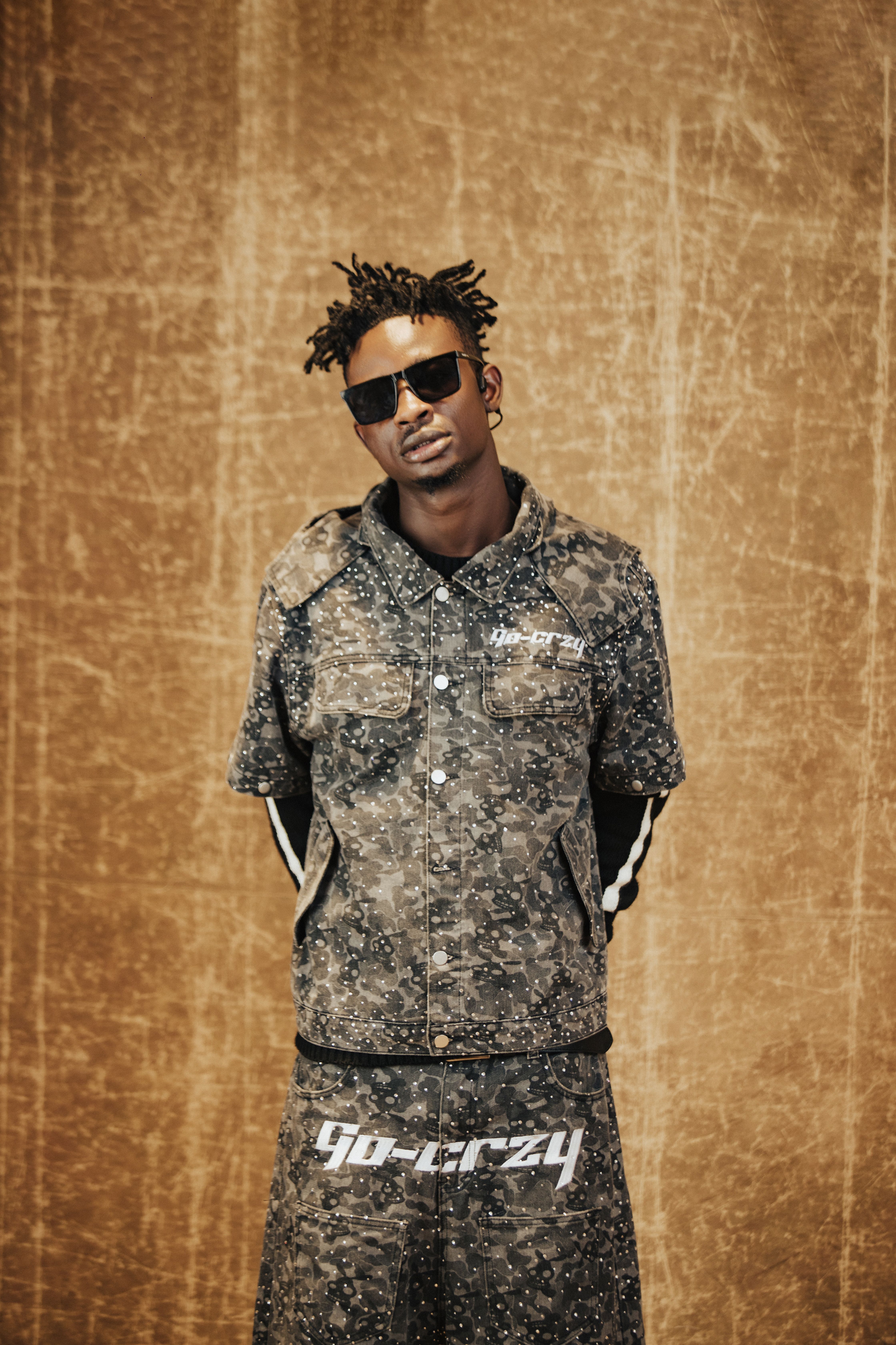 Fashionable man in camo outfit and sunglasses poses against a textured wall.