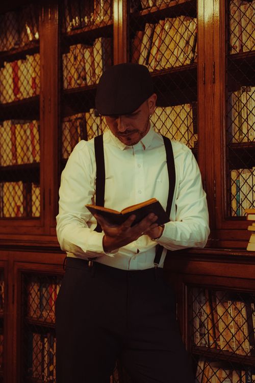 Free Adult man in vintage attire reading a book in a classic library setting. Stock Photo