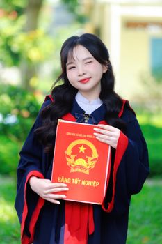 Smiling graduate in academic gown holding diploma on a sunny day.