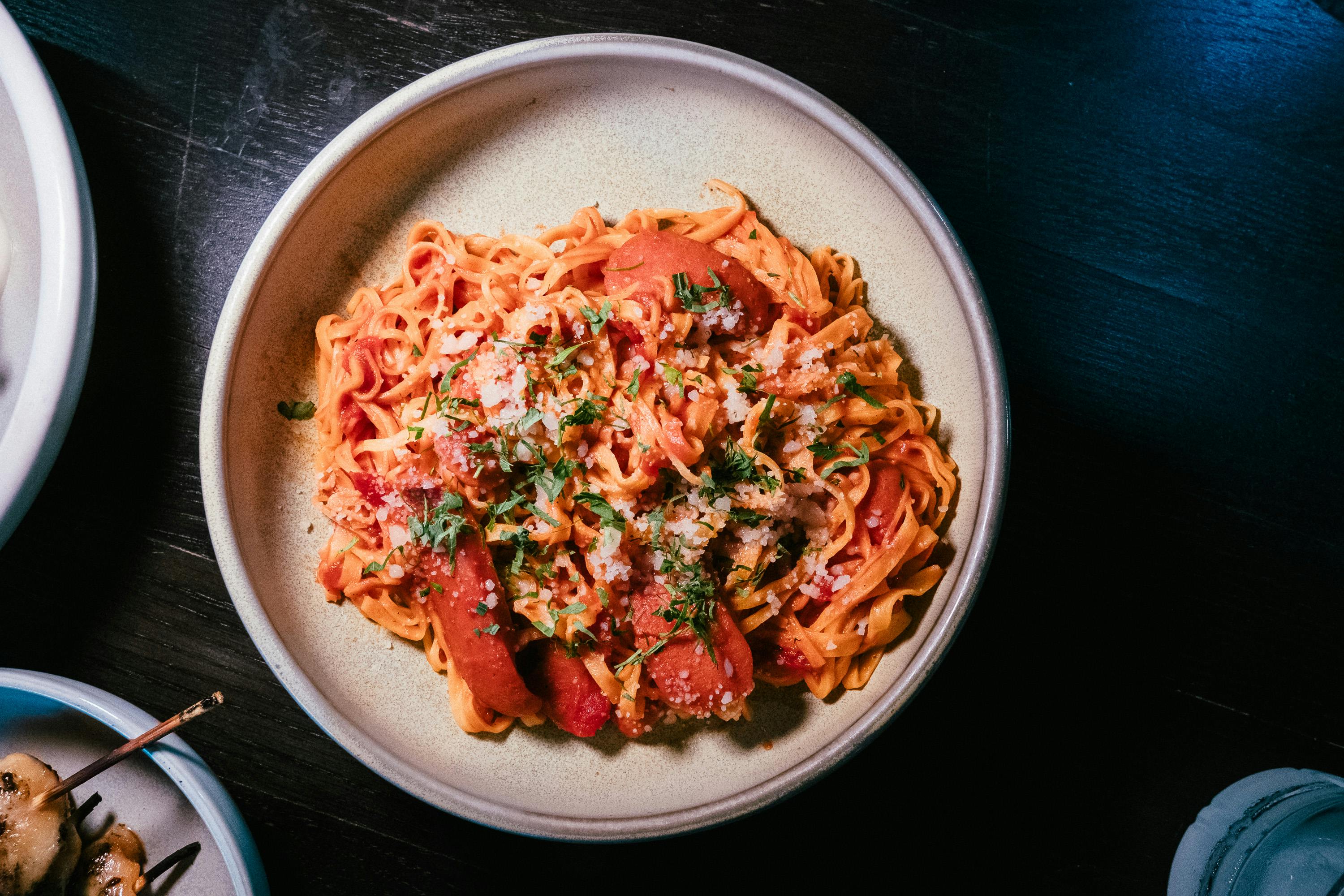 A steaming bowl of fresh Turkish noodles glossed with deep red pepper sauce, topped with crumbled white beyaz peynir, golden crispy garlic pieces, and bright green fresh parsley