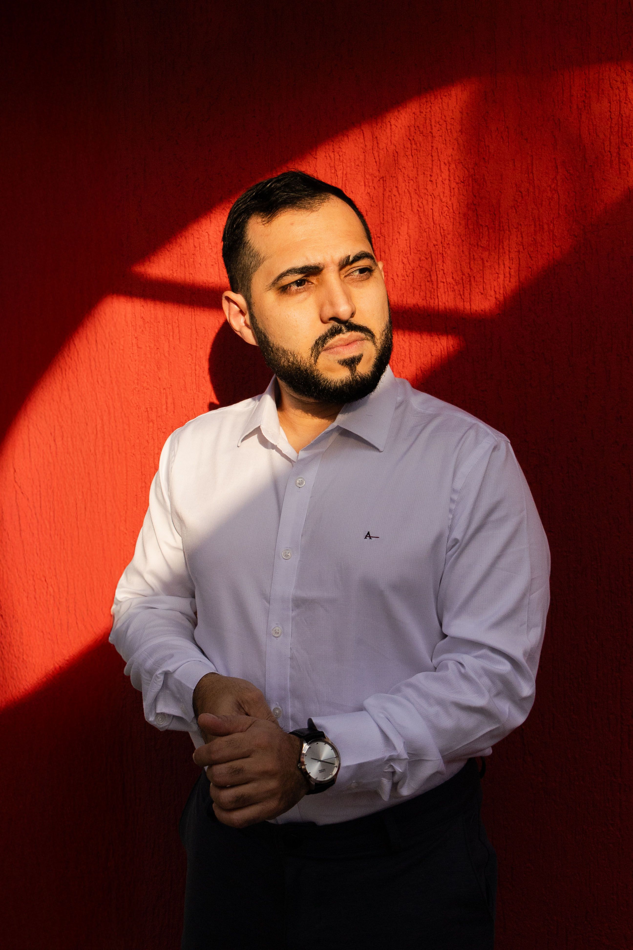 Stylish man in white shirt posing against a vivid red wall in sunlight.