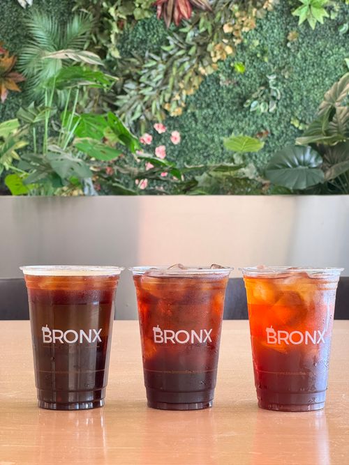 Free Three iced coffee cups labeled 'BRONX' on a table with lush green plants in the background. Stock Photo
