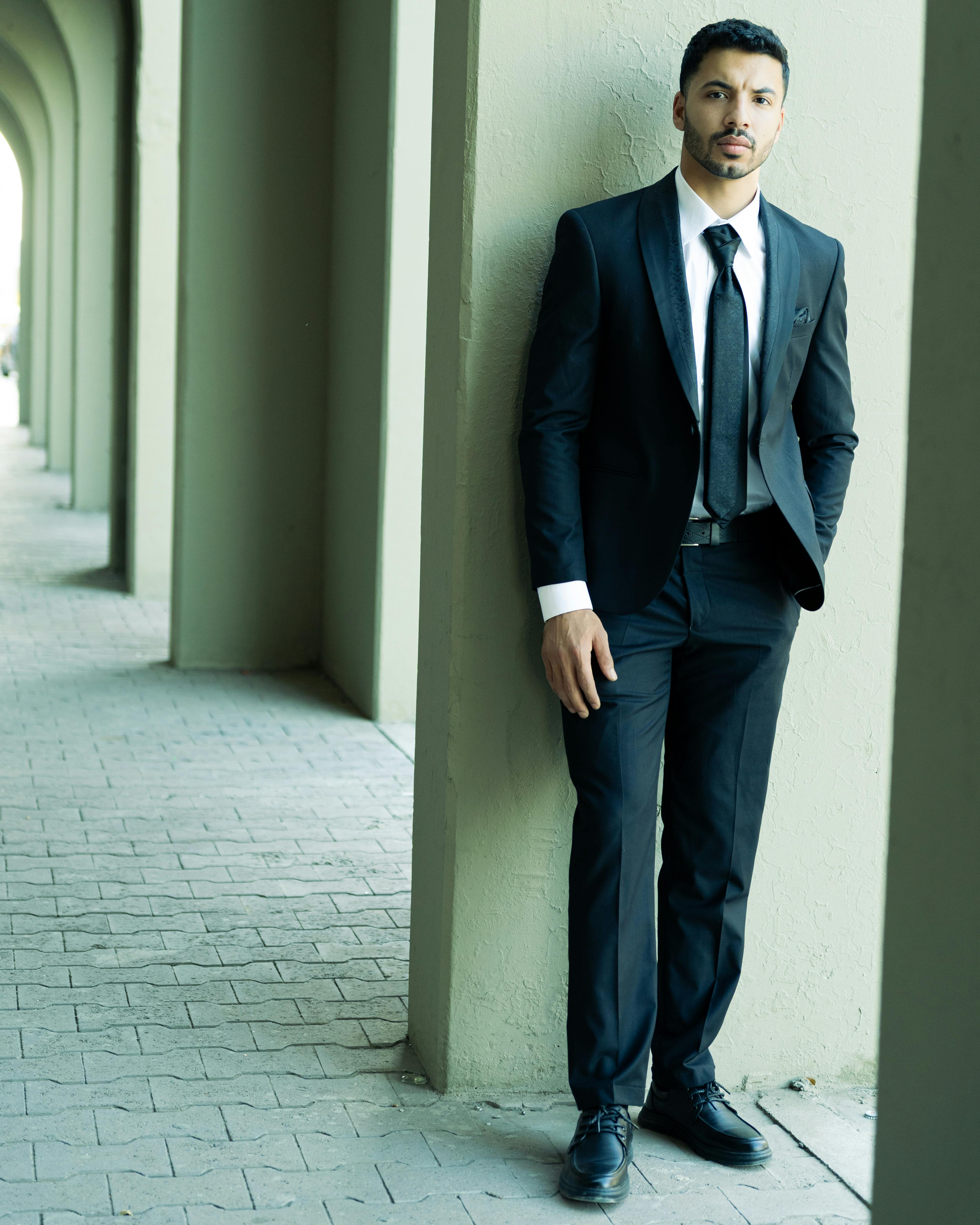 Stylish man in a black suit leaning against a wall in an architectural hallway. pmo manager training