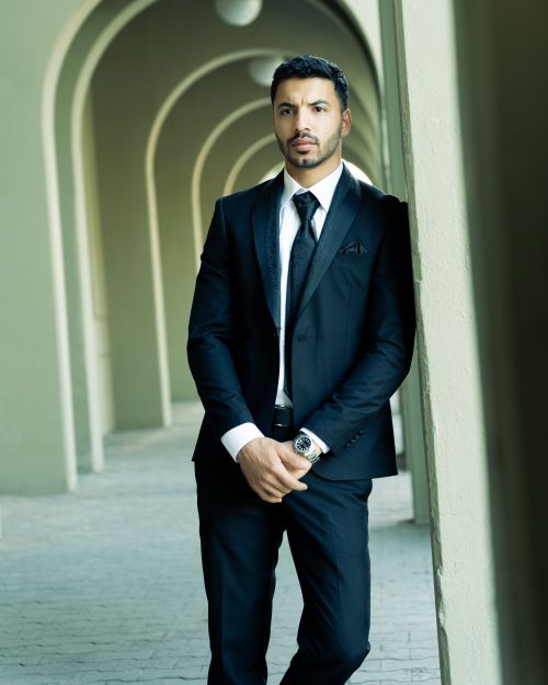 Free Young man in a sleek black suit leaning against a wall in an arched corridor. Stock Photo