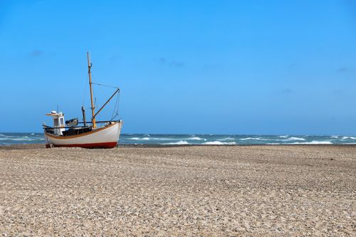 Free Scenic view of a wooden boat on a pebbly beach in Denmark under a bright blue sky. Stock Photo