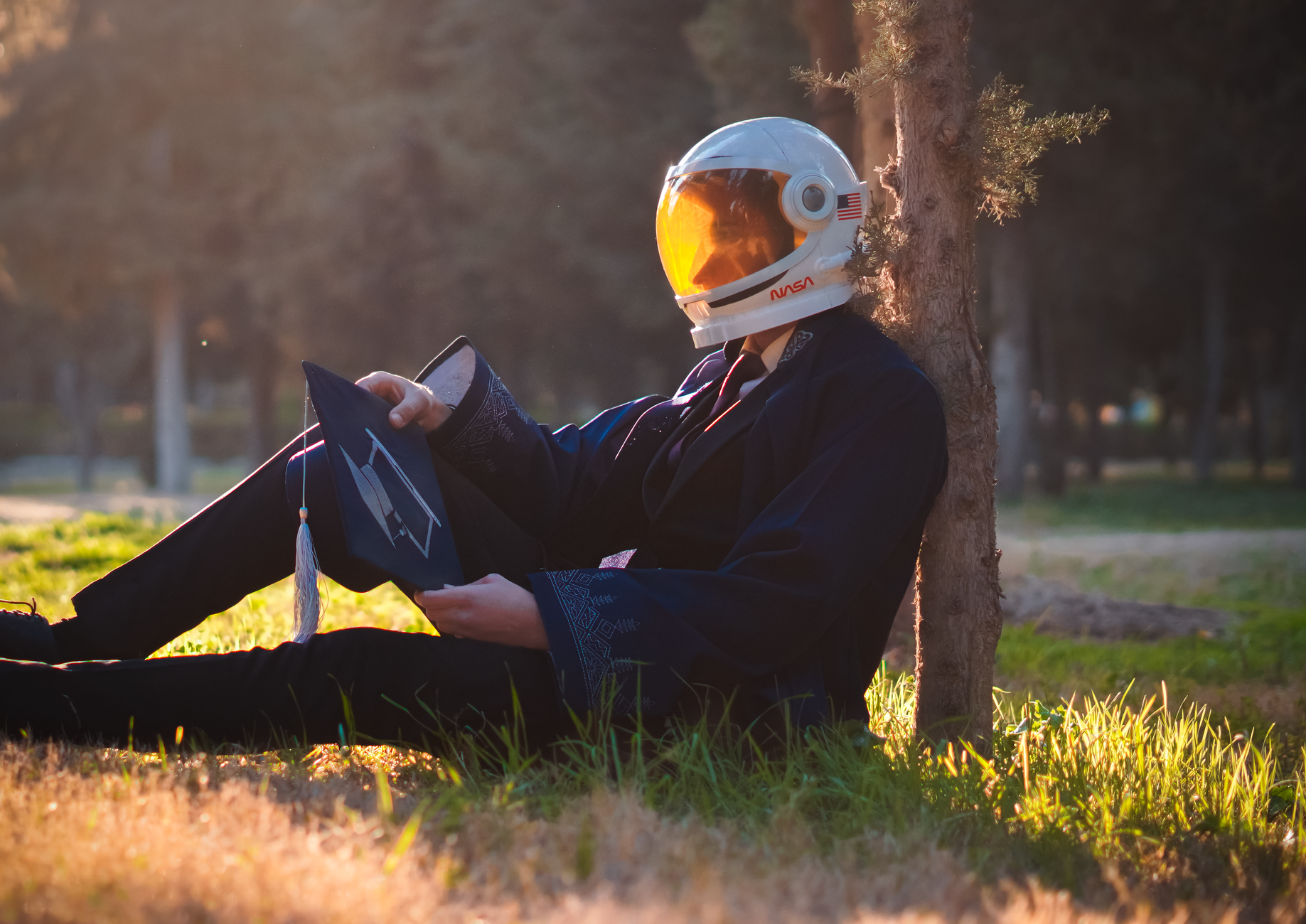 A person in astronaut gear and graduation robe sitting against a tree, symbolizing aspirations.