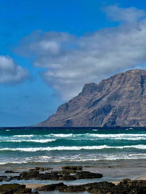 Free Dramatic ocean waves crash on the Lanzarote coastline with a rugged mountain backdrop under a vibrant blue sky. Stock Photo