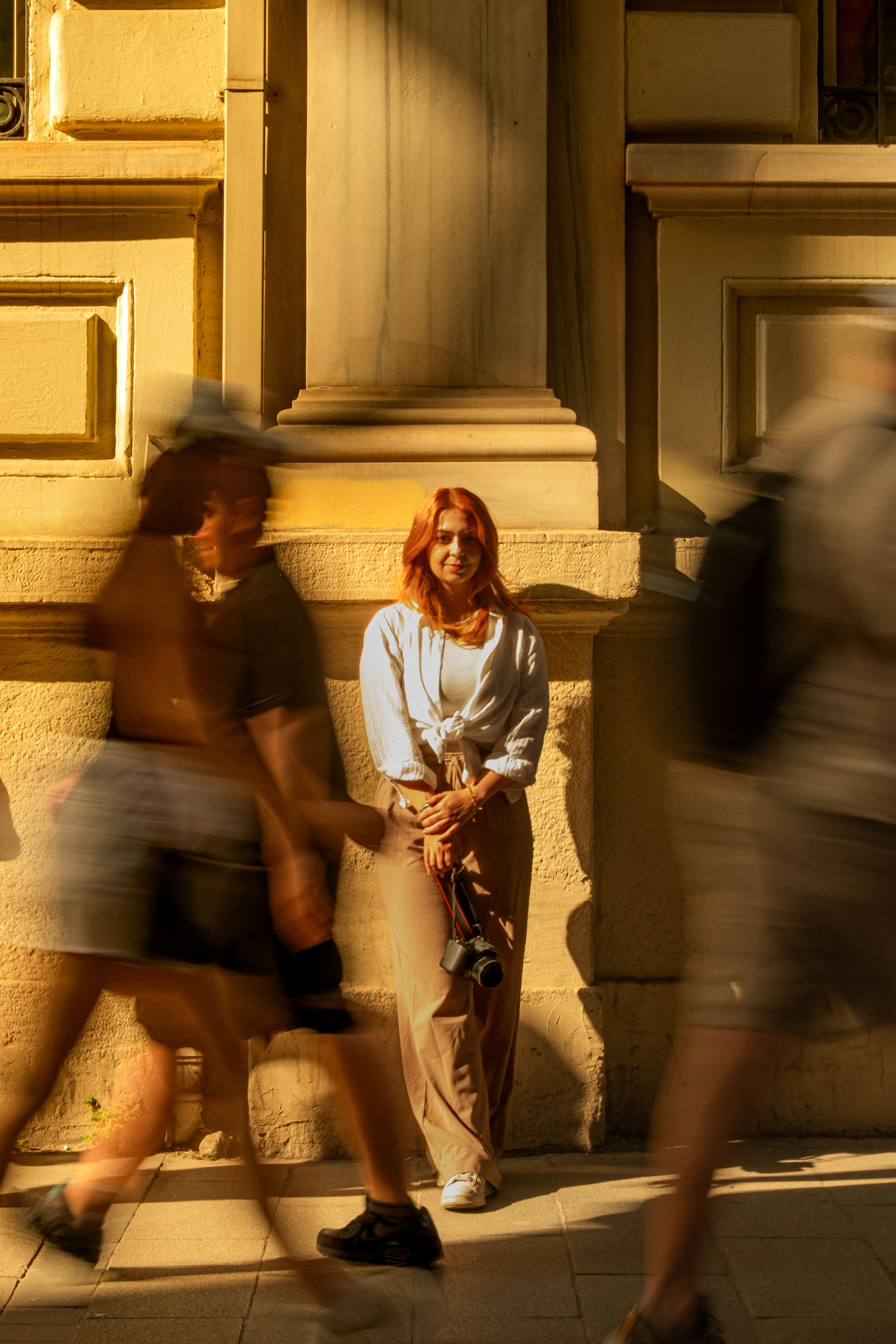 A young woman poses against a historic wall in İstanbul, Türkiye as people walk by.