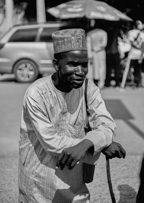 Free Black and white portrait of a man in traditional attire leaning on a cane, outdoors in bright daylight. Stock Photo