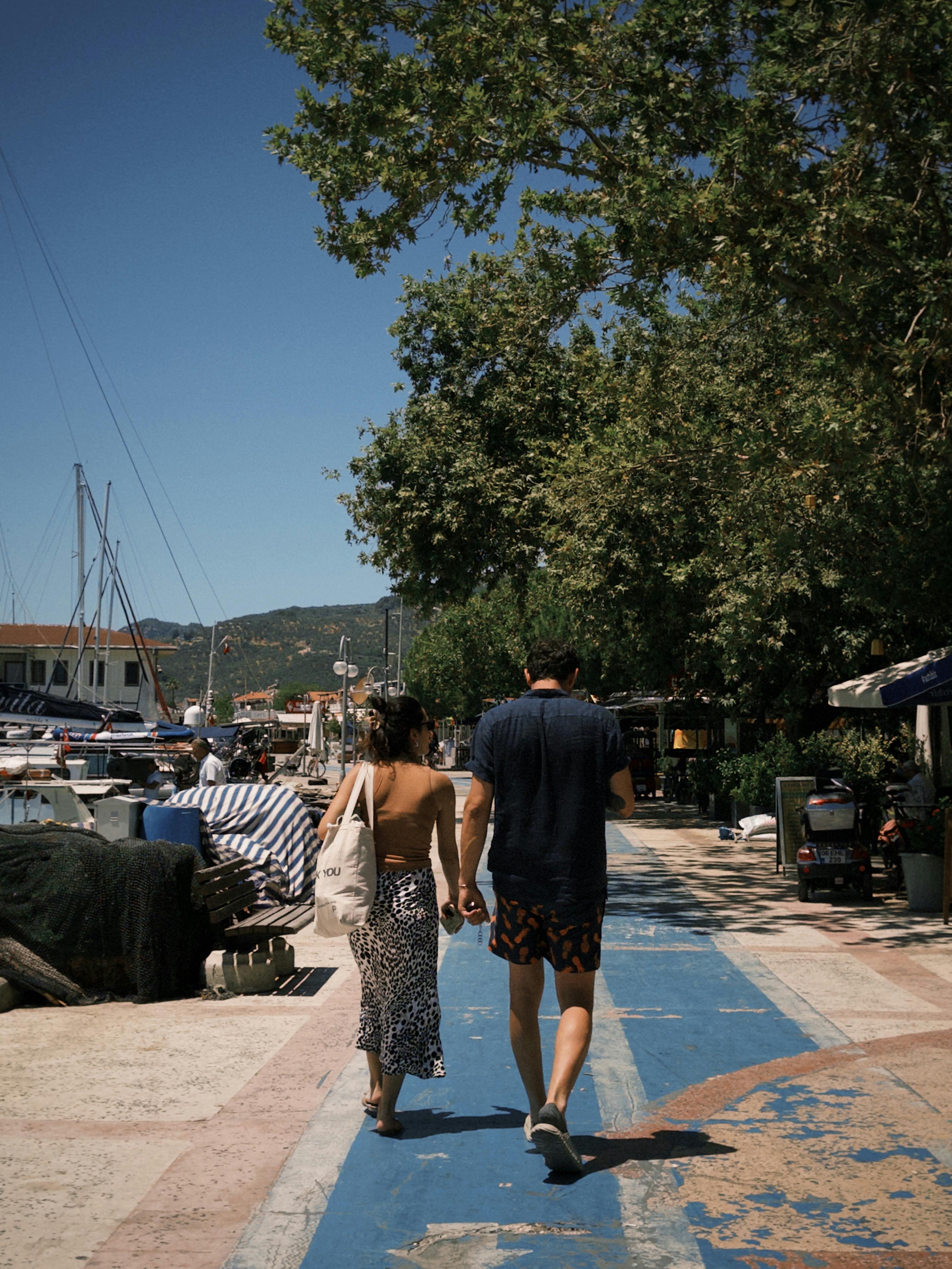 A couple walks along a sunny marina walkway lined with boats and trees, capturing a serene summer vibe.