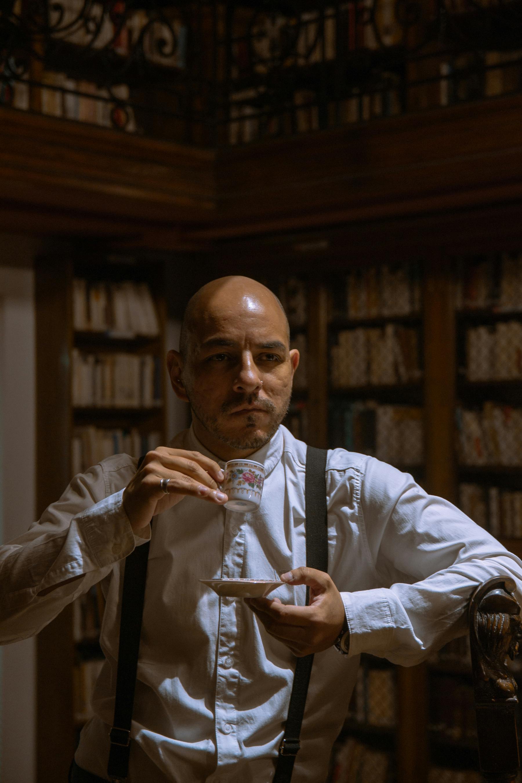 Bald man in formal attire enjoying coffee in a classic library setting.