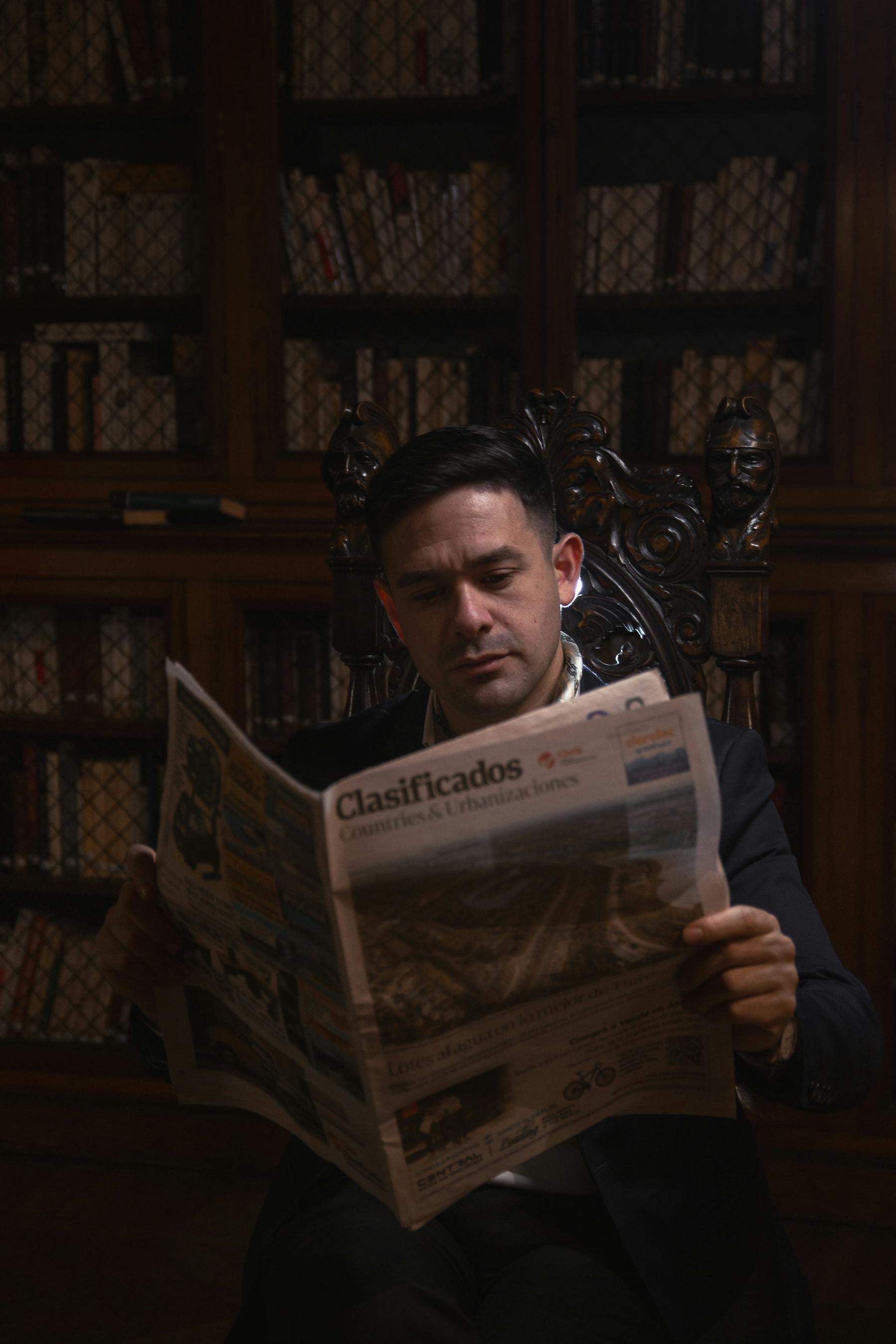 A man reading a newspaper while sitting in a dimly lit library with bookshelves in the background.