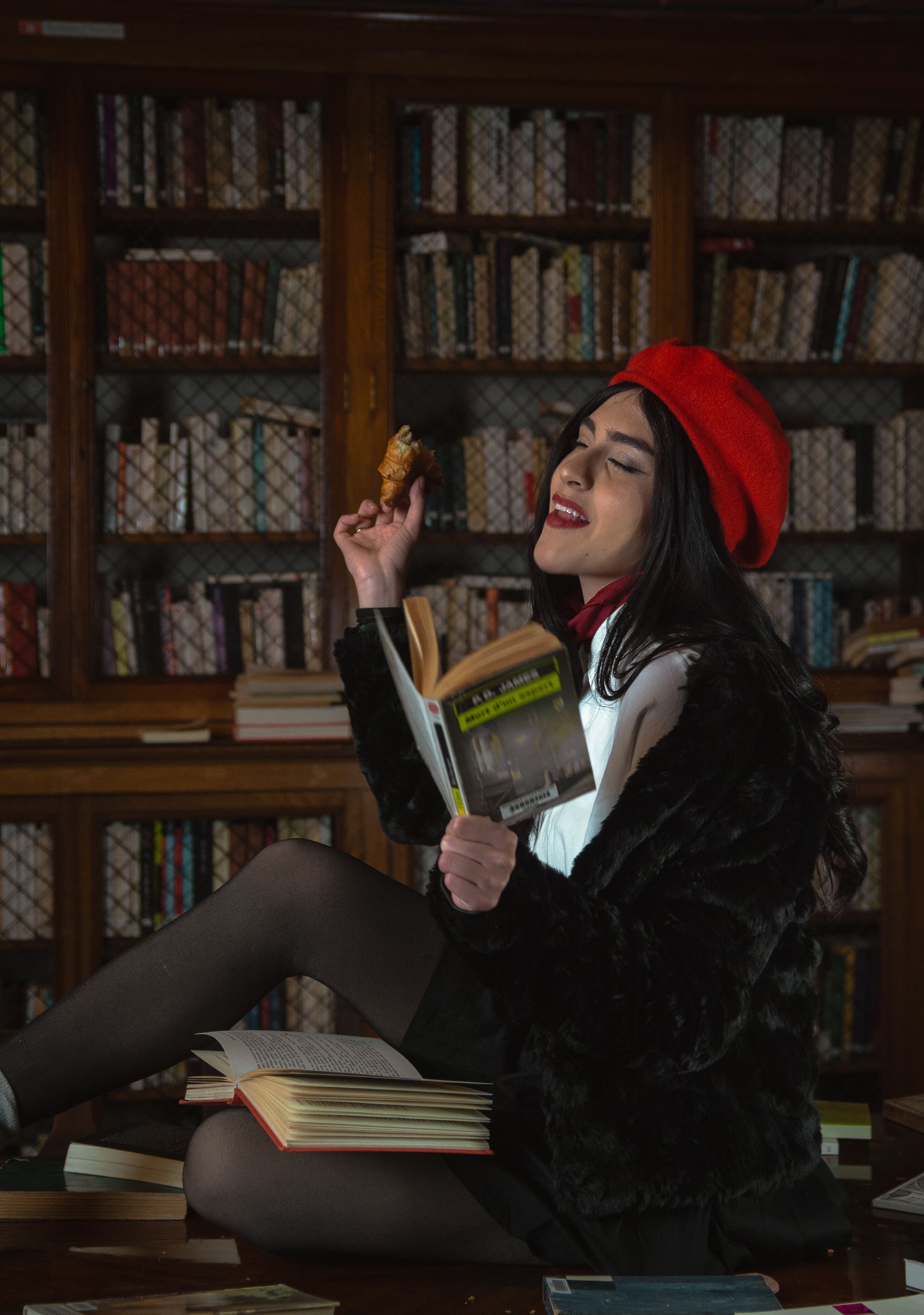 Gratis Una donna elegante con un basco rosso legge un libro e mangia un pasticcino in un'accogliente biblioteca. Foto a disposizione
