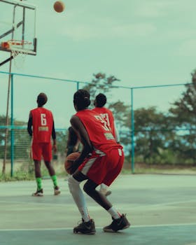 Teenagers playing basketball on an outdoor court, focusing on a player in a red jersey preparing to shoot.