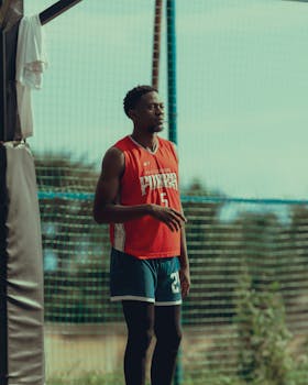 Focused basketball player in a red jersey stands on an outdoor court during the day.