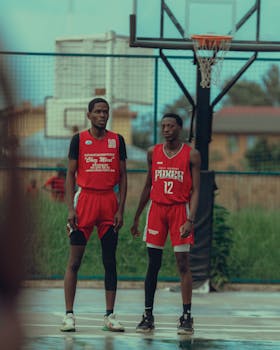 Two young basketball players in red uniforms standing on an outdoor court under a cloudy sky.