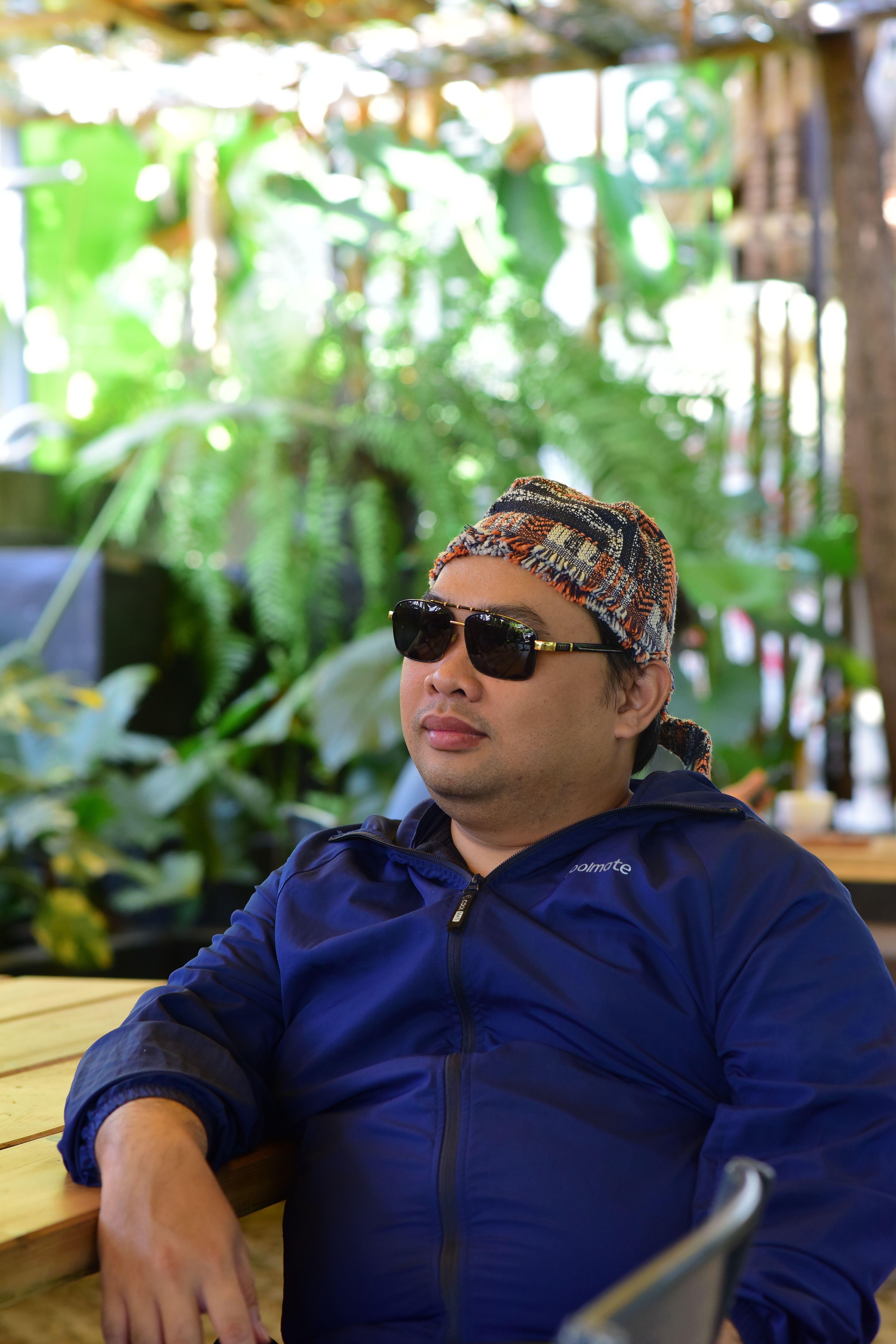 Man in sunglasses and traditional headwear relaxing outdoors with greenery backdrop.