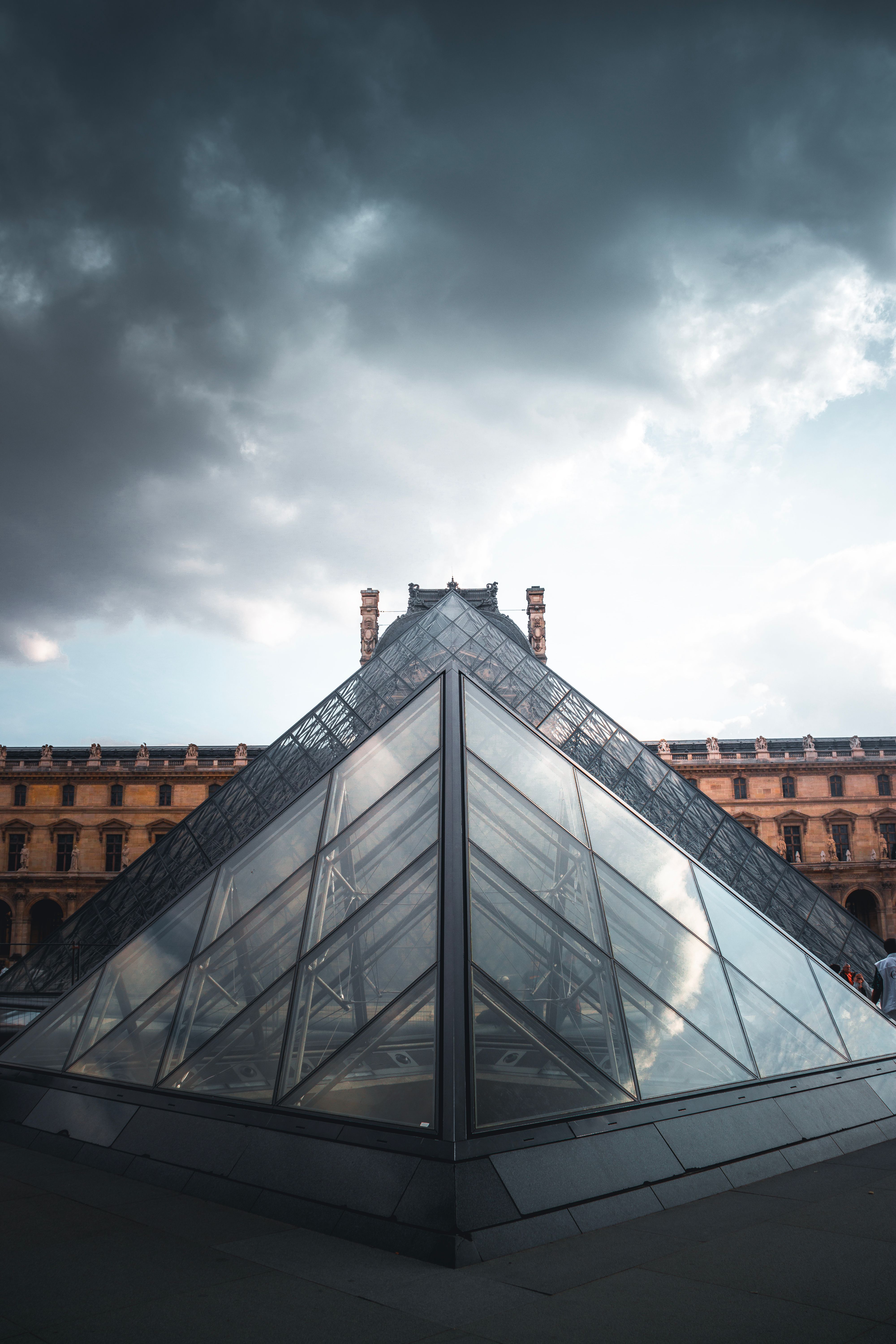 The glass Louvre Pyramid in Paris under a dramatic cloudy sky, a real stock photo with no people in the frame that sets the scene for Lupin Part 4, where simultaneous museum heists put Assane Diop's name back in the headlines