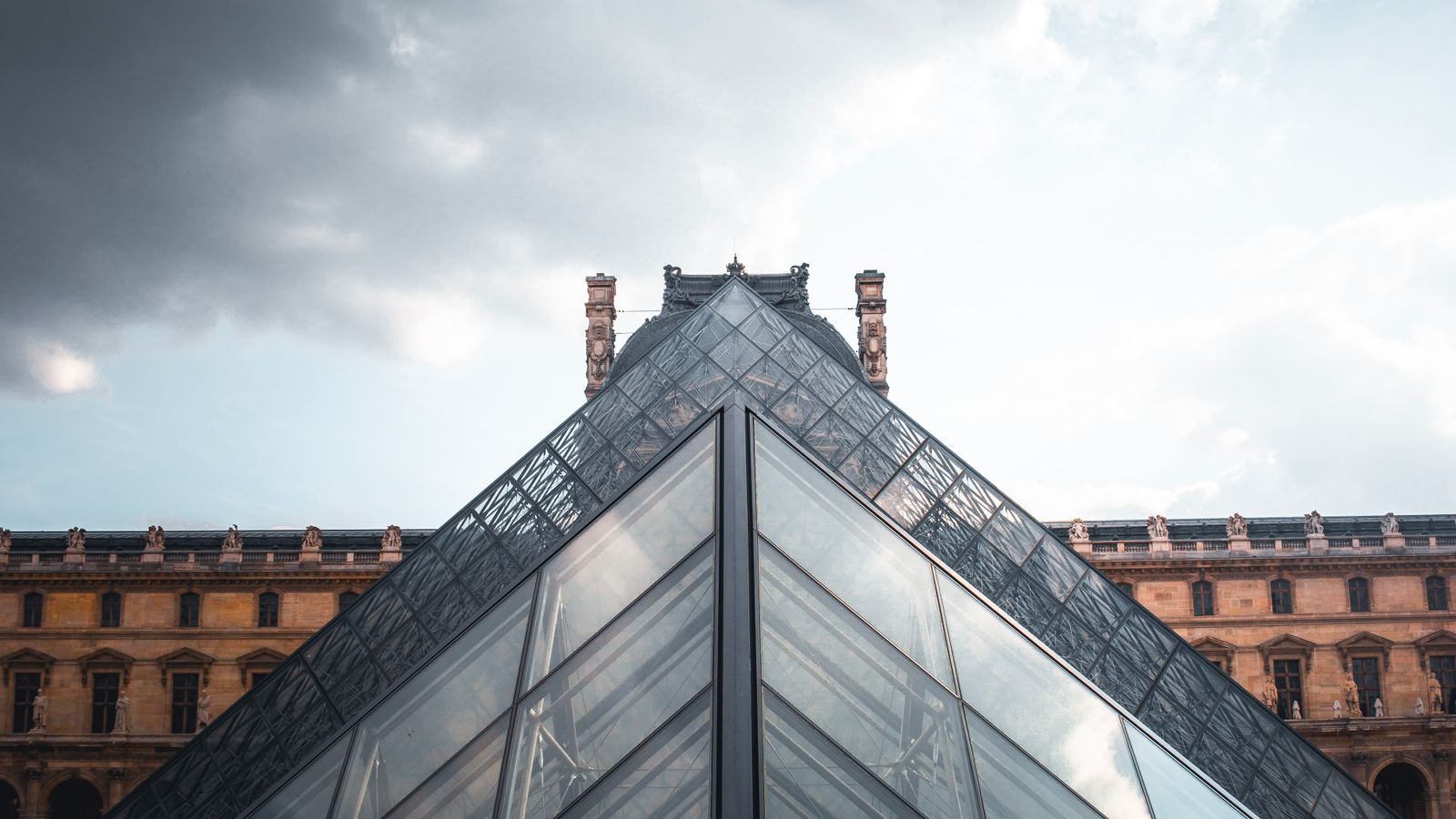 The glass Louvre Pyramid in Paris under a dramatic cloudy sky, a real stock photo with no people in the frame that sets the scene for Lupin Part 4, where simultaneous museum heists put Assane Diop's name back in the headlines