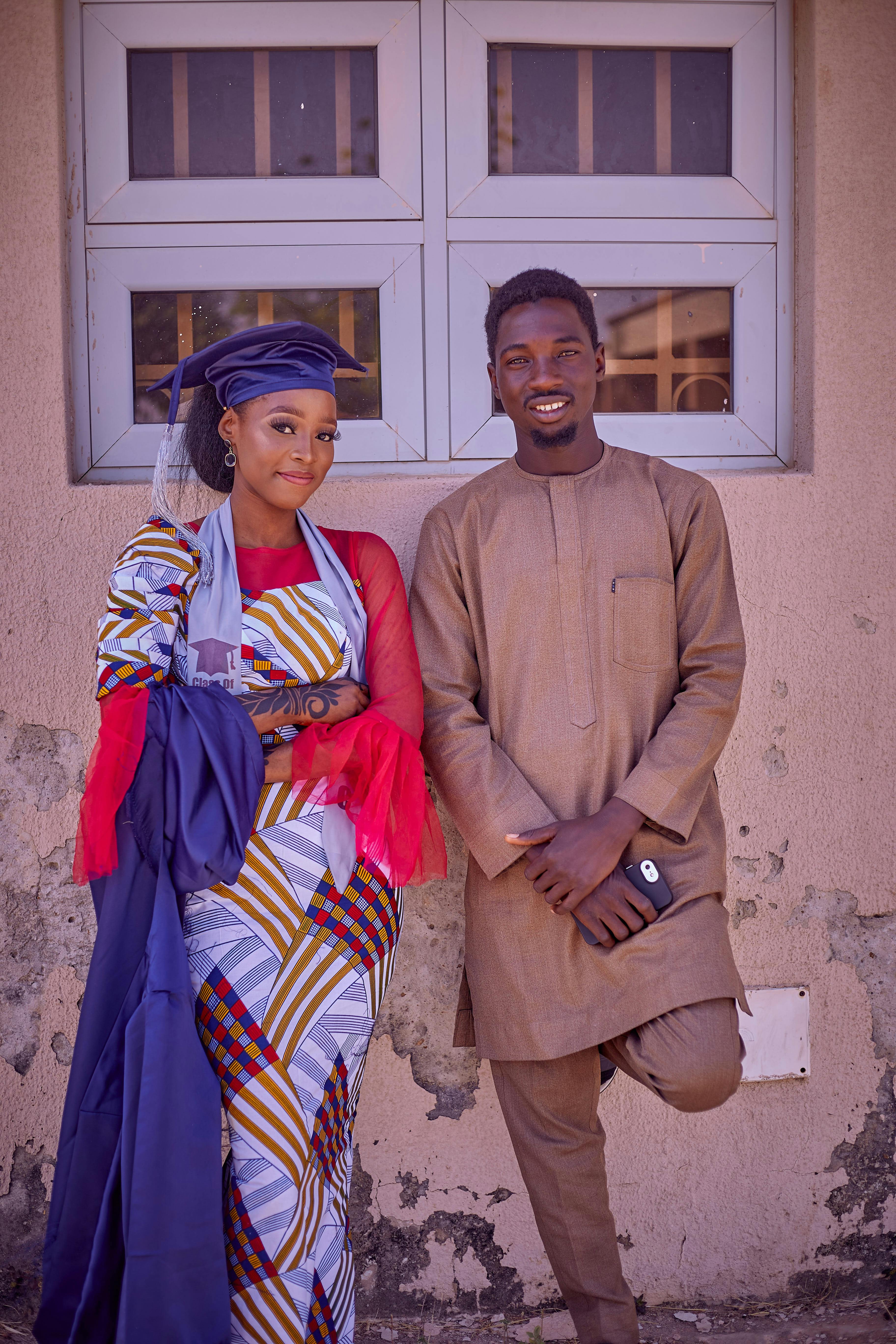 A couple dressed in vibrant traditional African garments poses outside a window.