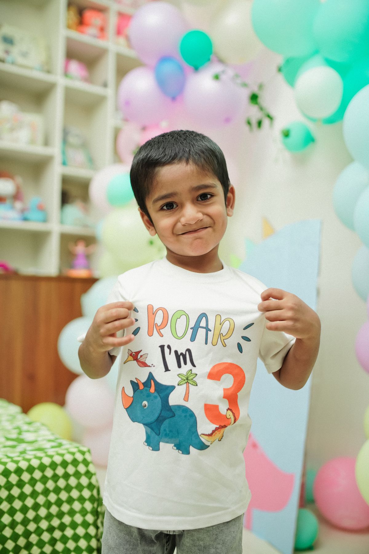 Adorable child celebrating a dinosaur-themed third birthday party indoors with balloons. Stock Photo.