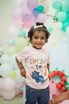 Cute child with a dinosaur-themed shirt celebrating their 3rd birthday with colorful balloons indoors.