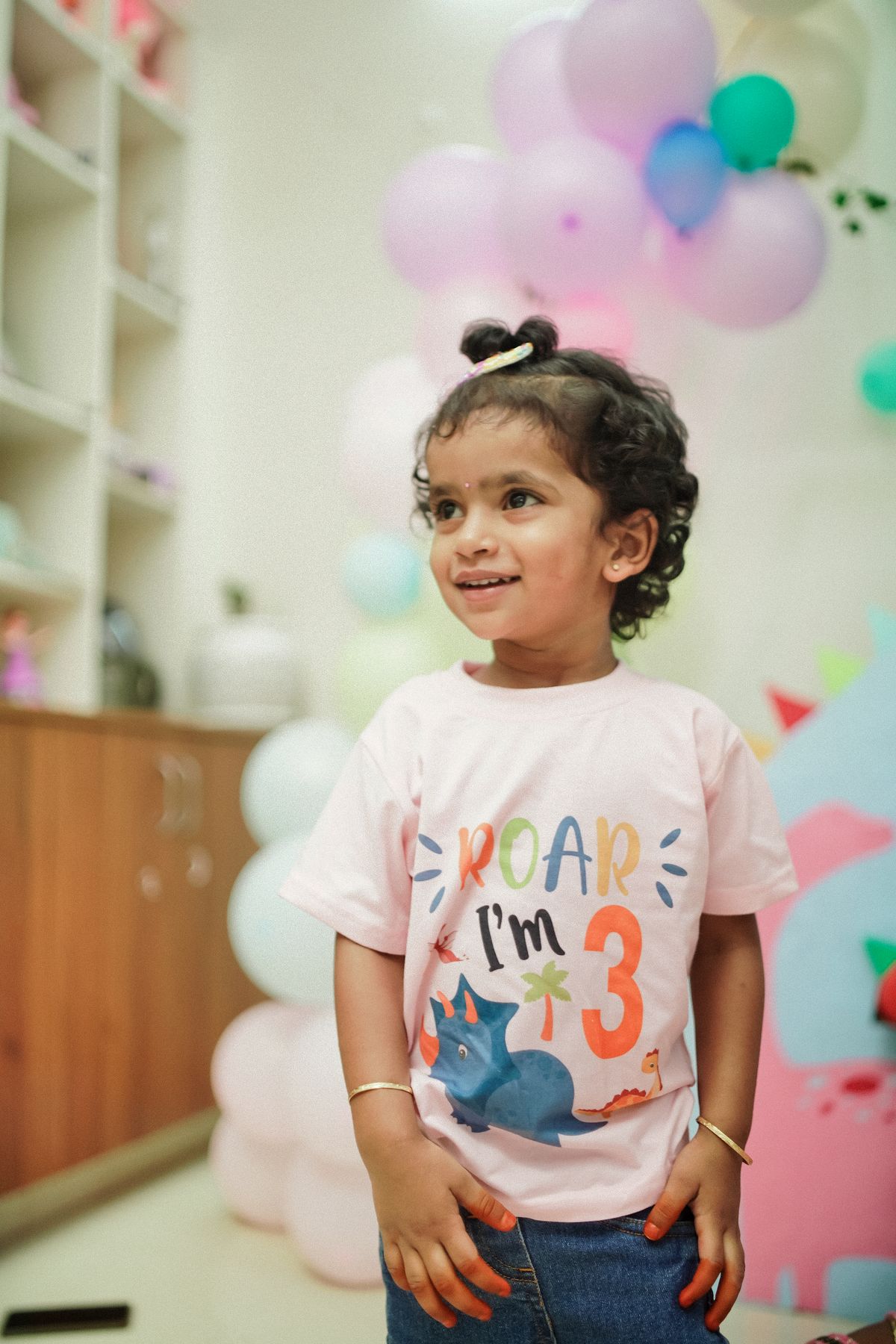 Smiling toddler celebrating a dinosaur-themed third birthday indoors. Stock Photo.