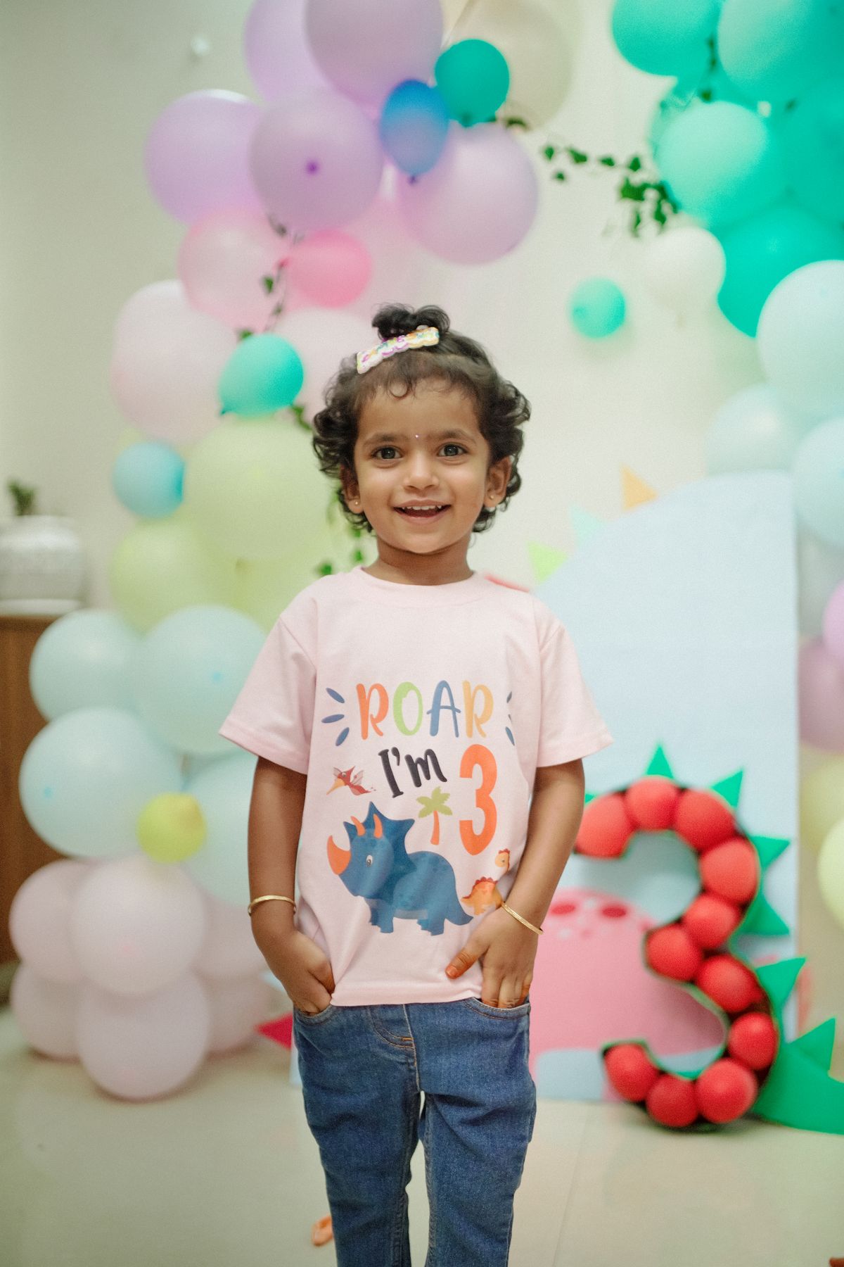 Cute child celebrating a third birthday with a dinosaur theme and colorful balloons. Stock Photo.