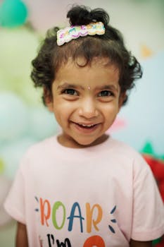 A young girl smiling brightly at her dinosaur-themed birthday party indoors with colorful balloons.