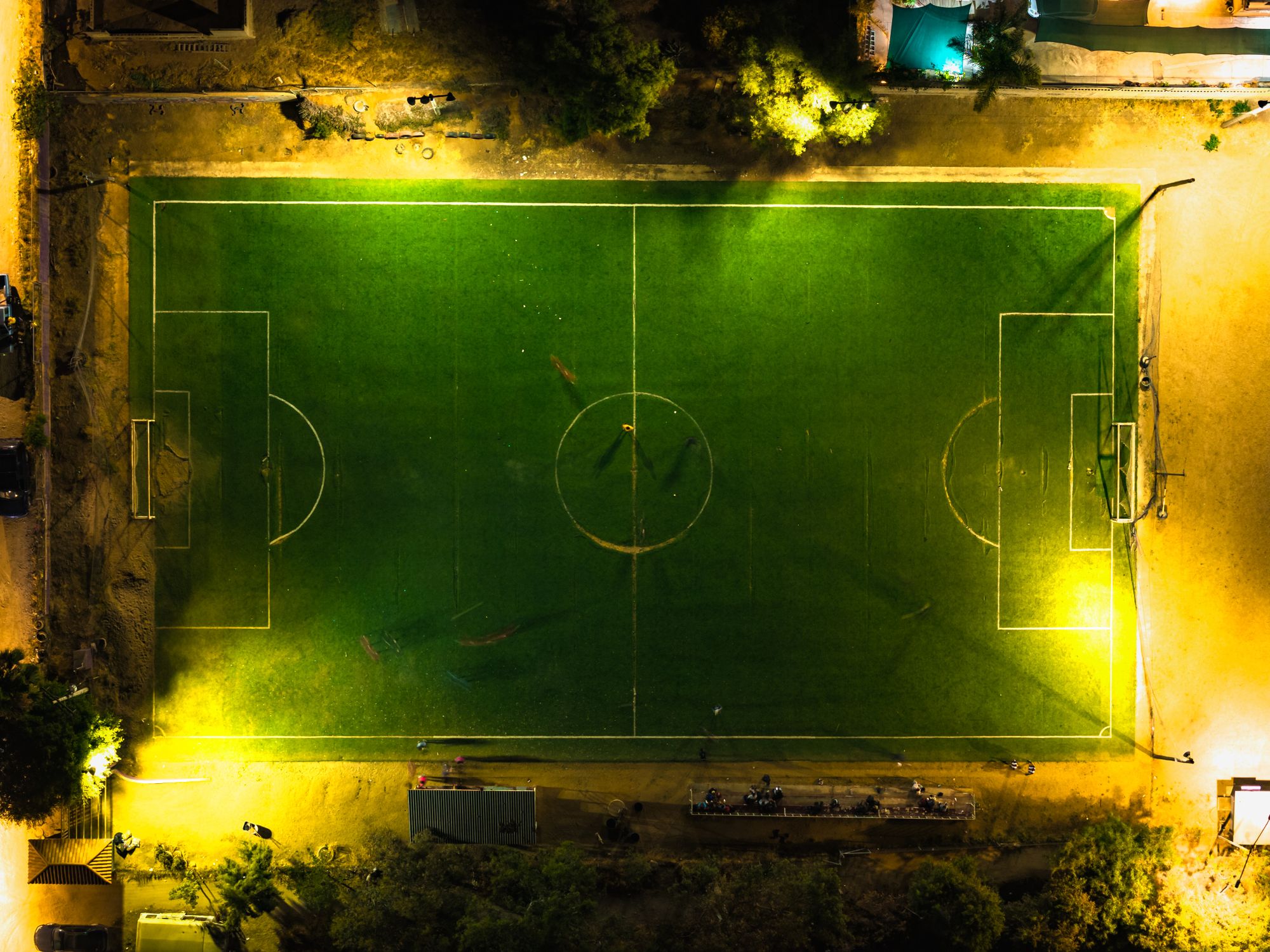 Grada de un estadio de fútbol modesto con aficionados bajo los reflectores al anochecer