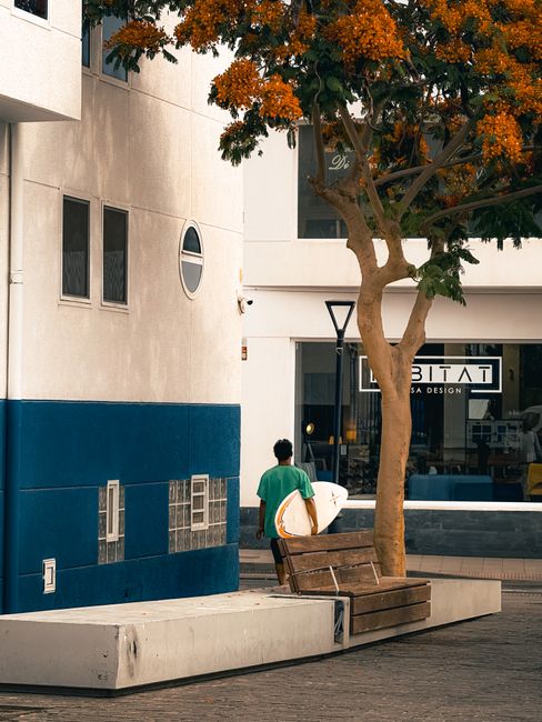 A surfer walks through a city street with a surfboard, showcasing the blend of urban and surf culture in the Canary Islands.